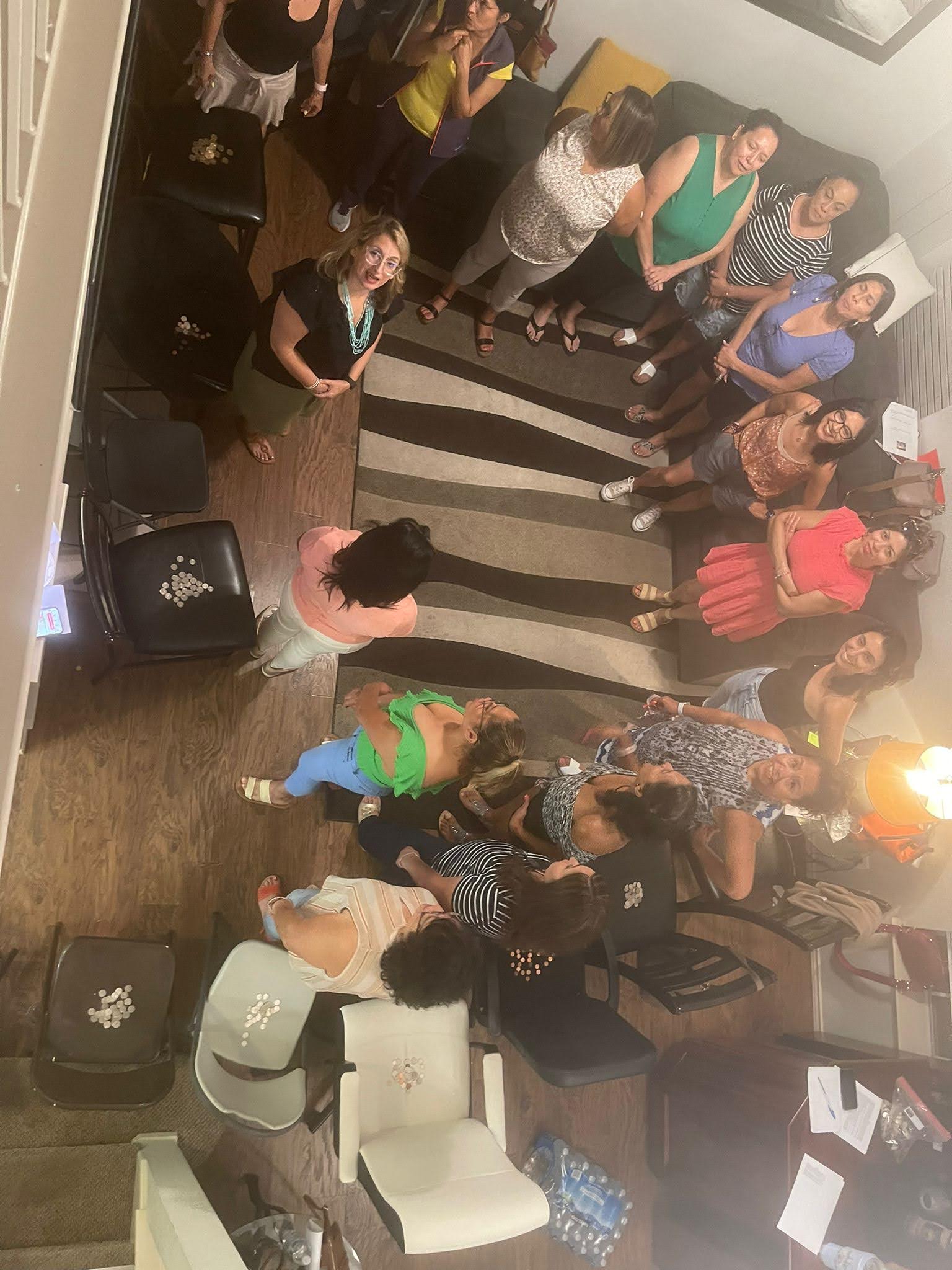 Group of people gathered in a room, some standing and some sitting, with a white chair and a wooden floor.