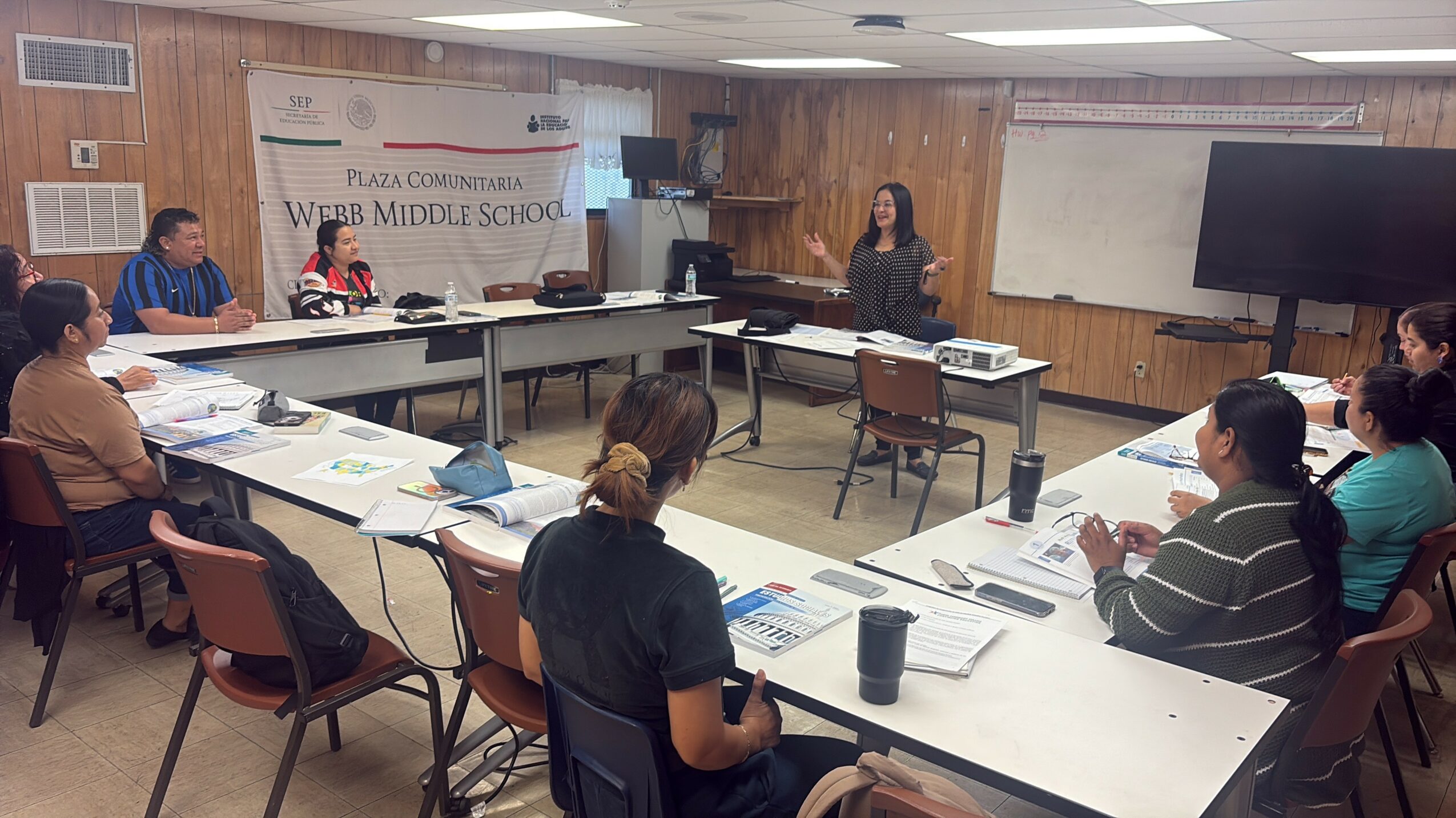 People seated around a U-shaped table in a classroom, with a woman standing at the front, presenting. Whiteboard and TV screen visible.