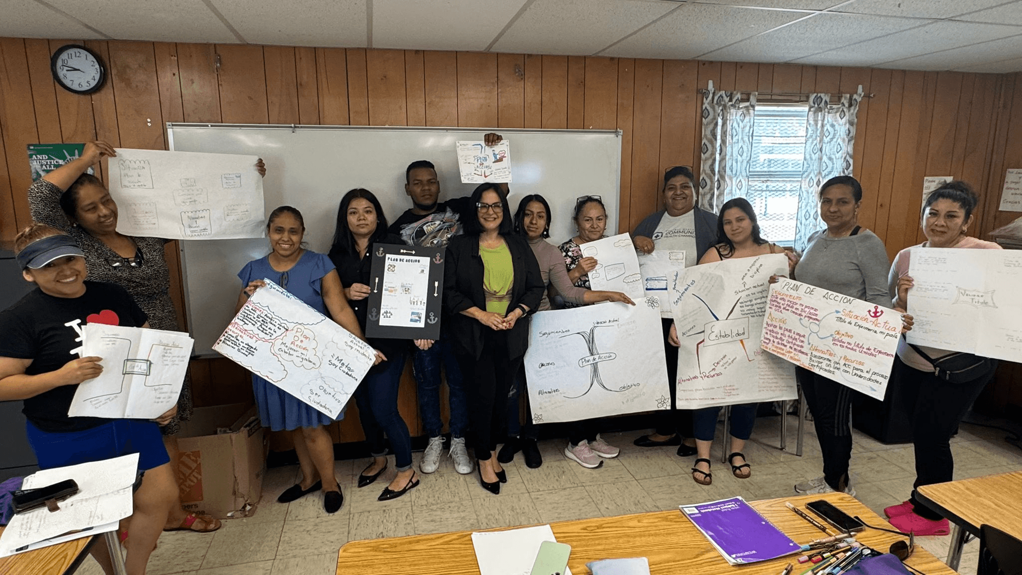 Group of people holding posters and a tablet in a classroom with whiteboard and window, wooden walls, and a clock.