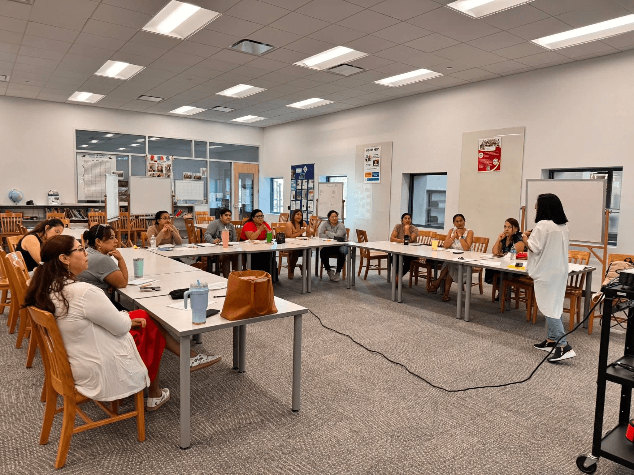 Group of people seated around tables in a classroom or meeting room, listening to a person standing and speaking.