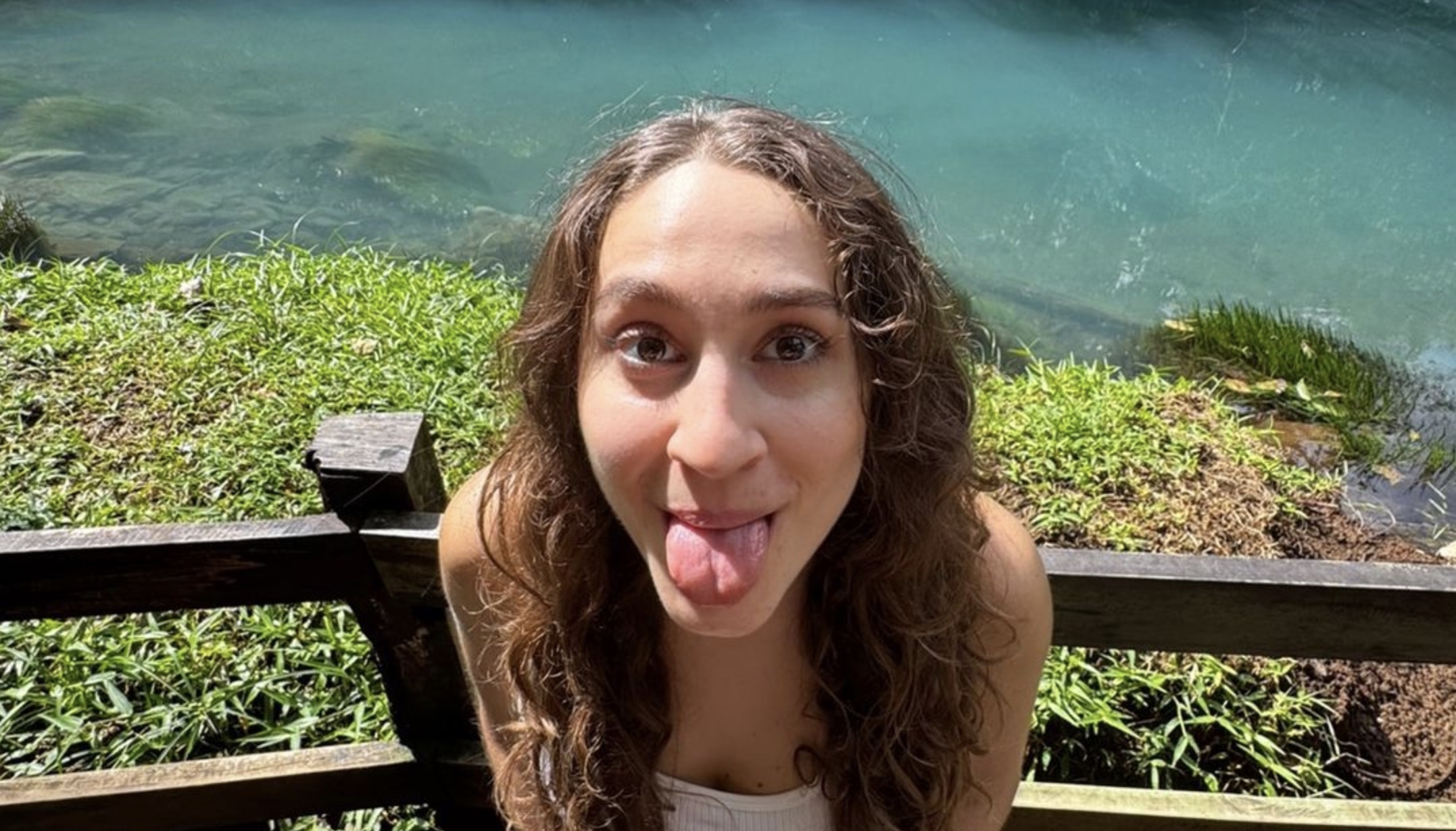 Young woman with curly hair sticking out her tongue outdoors near water, smiling, with a wooden railing behind her.