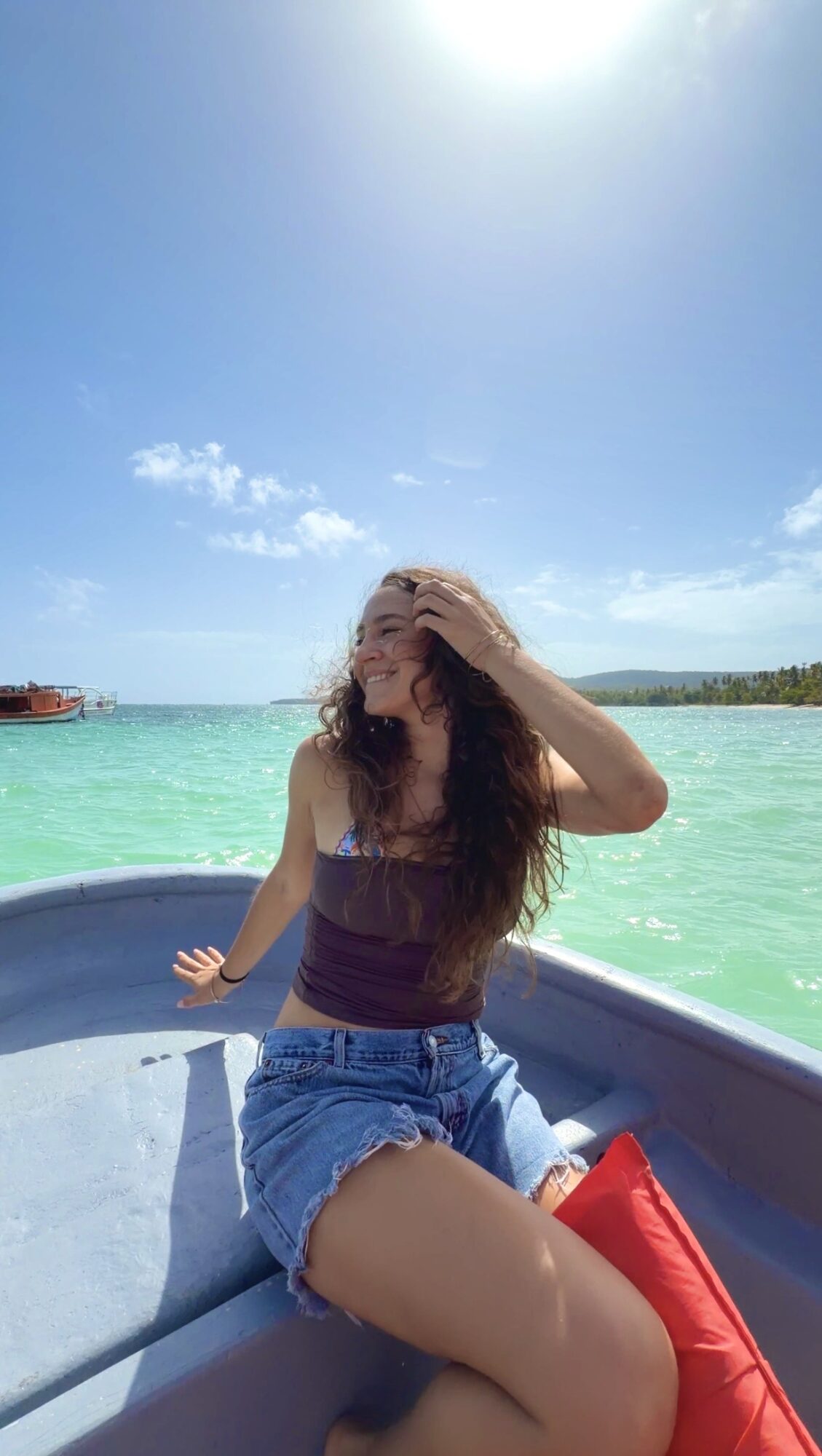 Young woman sitting on a boat with turquoise water and blue sky, smiling and touching her hair, sunlight overhead.