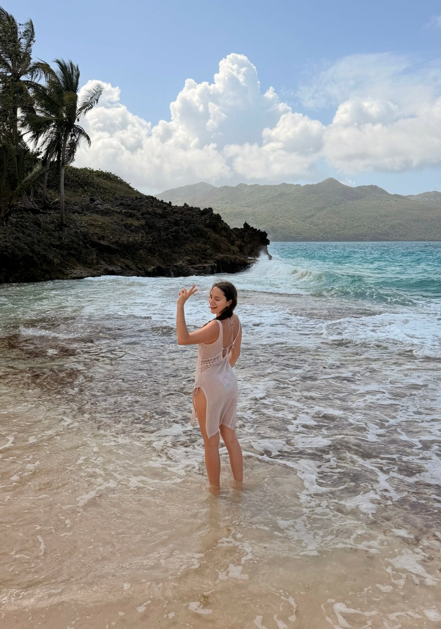 Woman standing in shallow ocean water, waving, with a tropical beach, palm trees, and mountains in background.