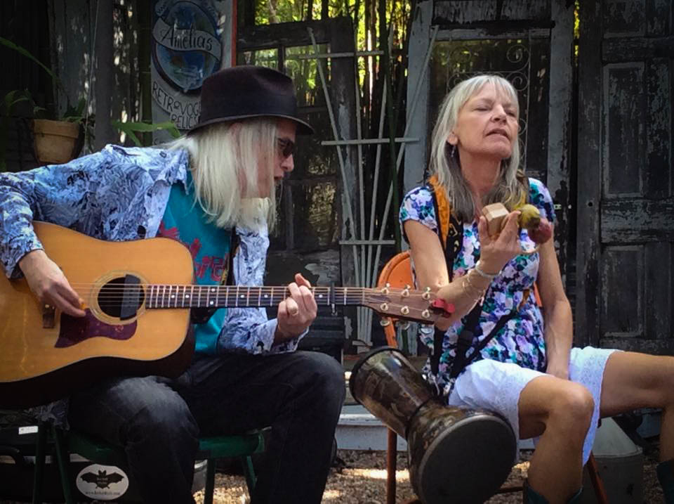 Two women sitting outdoors, one playing guitar and the other holding a small object, with a wooden fence in the background.