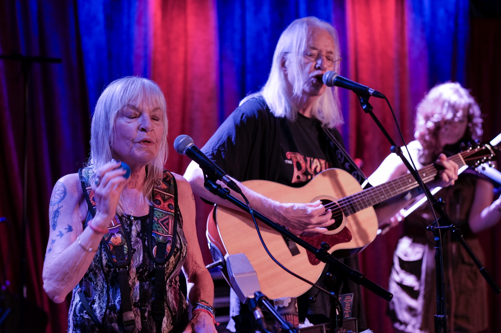 Three older women performing on stage with microphones and a guitar, colorful curtains in the background.