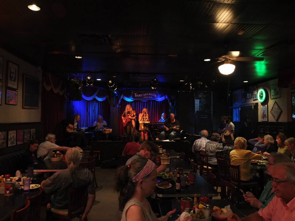 People seated at tables watching a live band on stage in a dimly lit venue with blue curtains and colorful lighting.