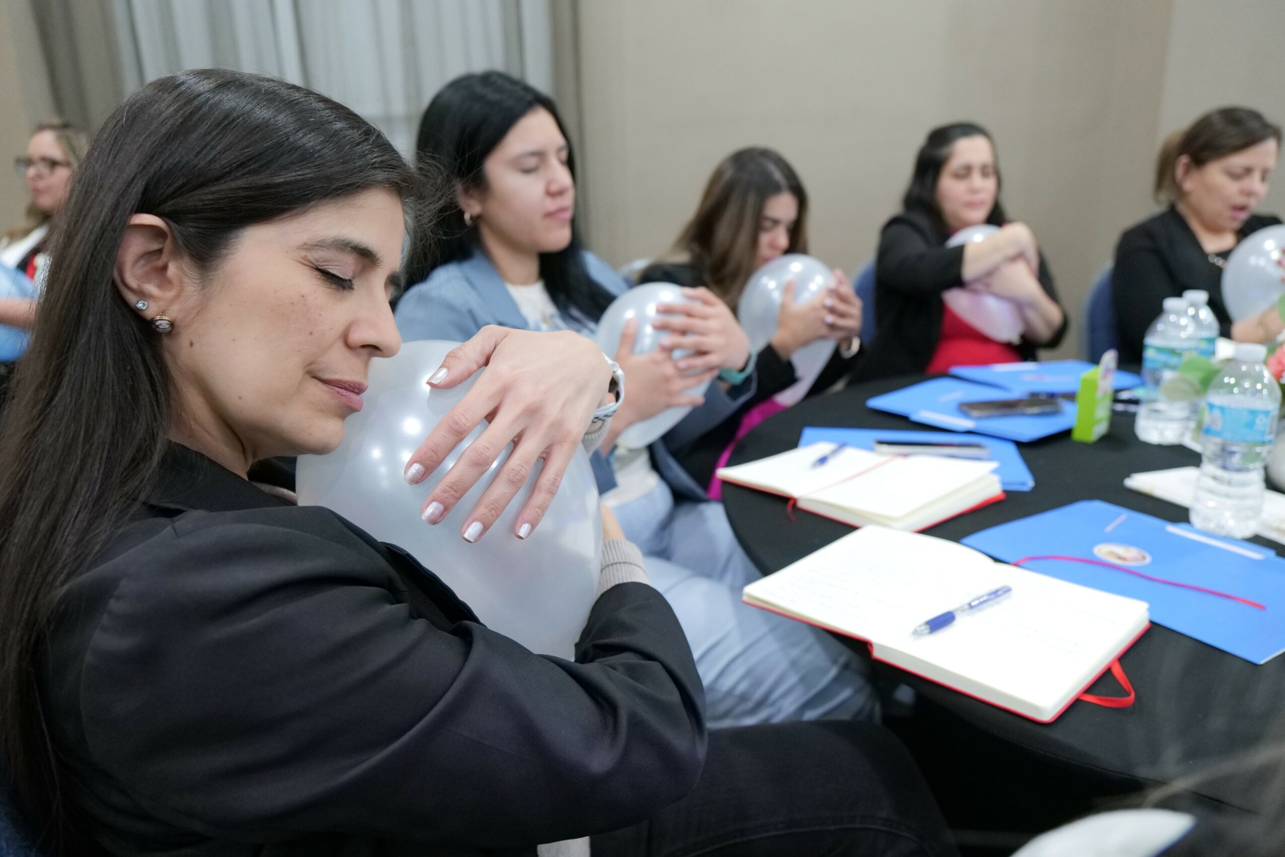 Group of women sitting at a table, holding clear plastic bags, with notebooks and water bottles in front, in a meeting room.