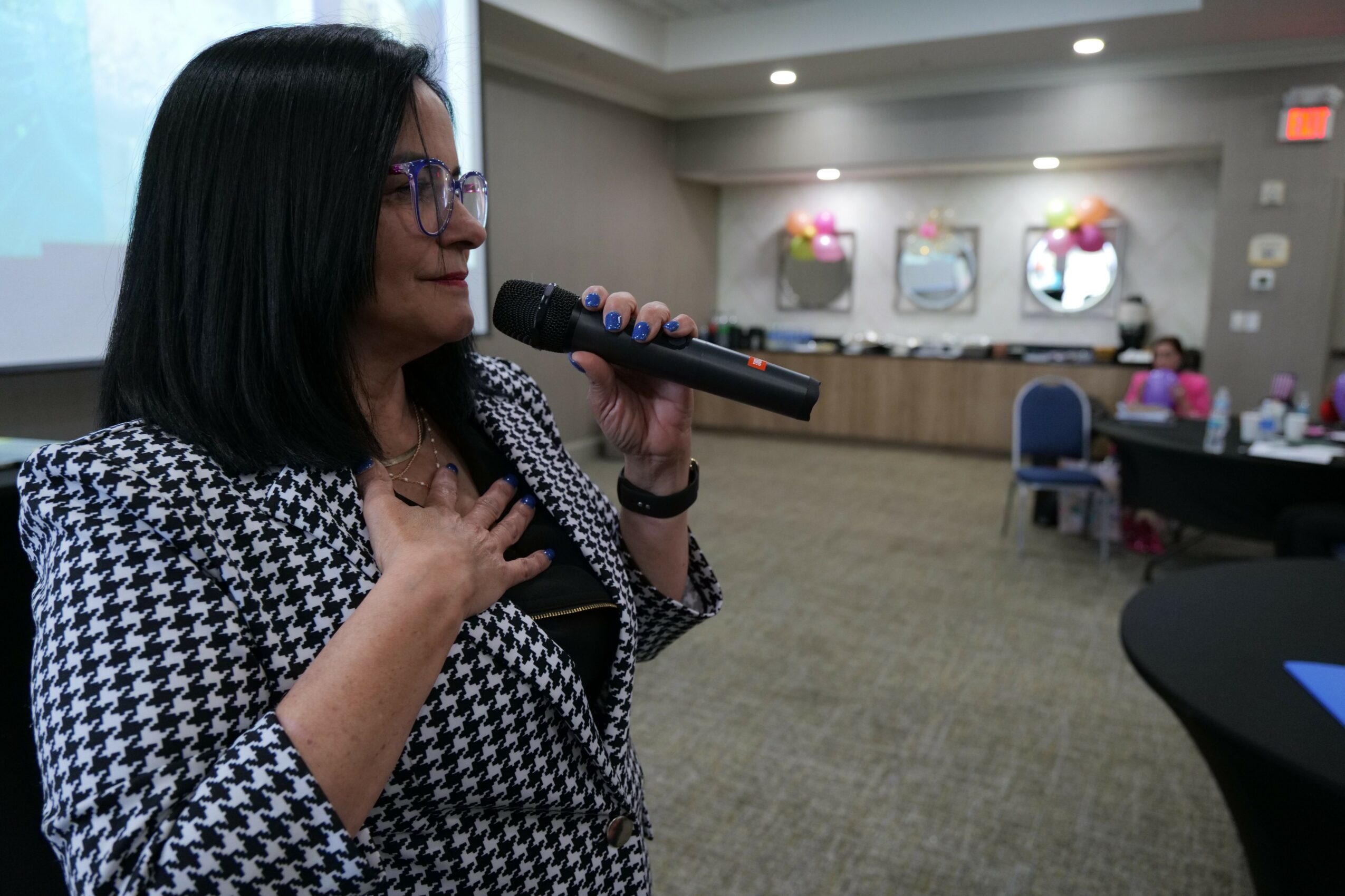 Woman speaking into a microphone in a conference room with tables and chairs, decorated with flowers on the wall.