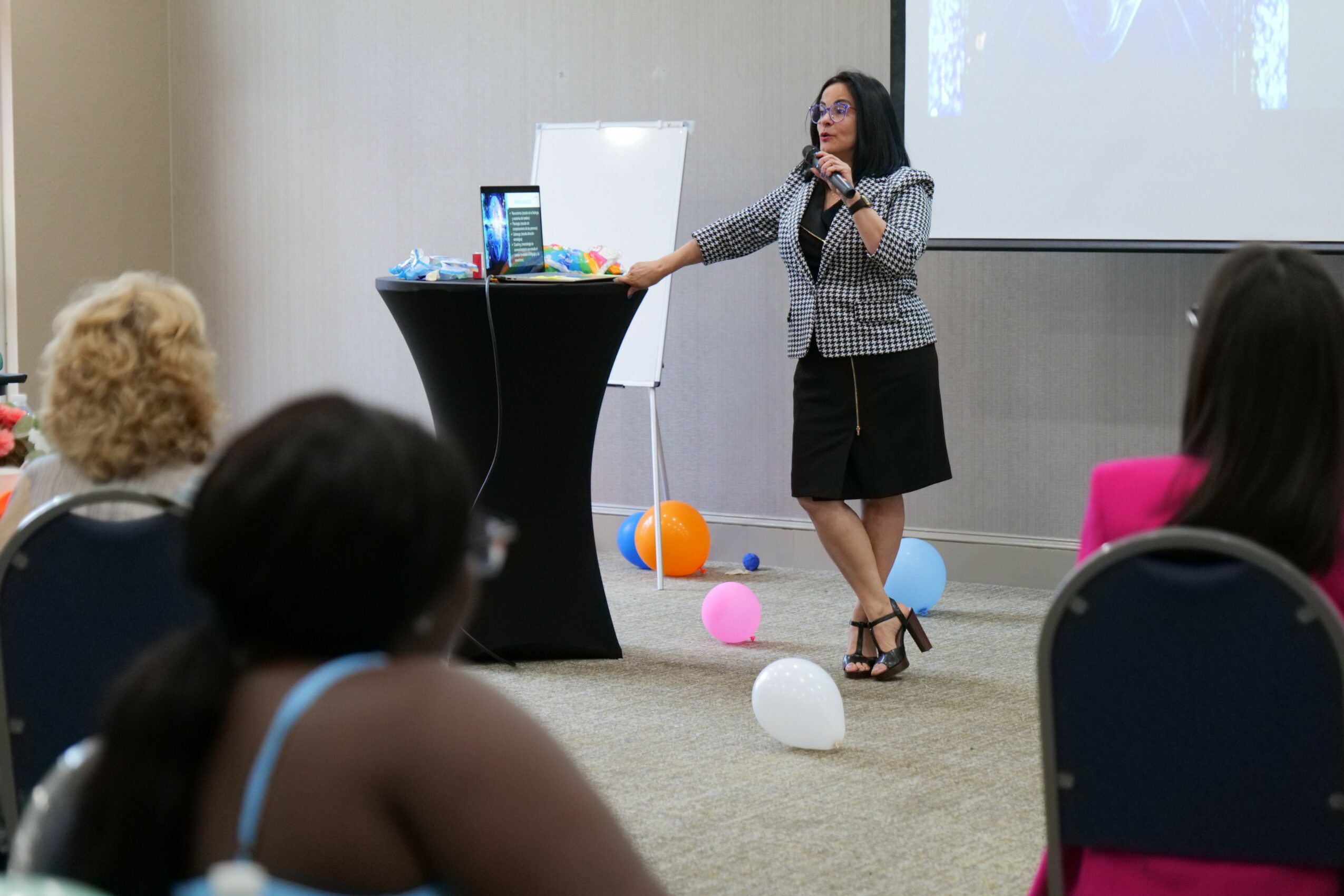 Woman speaking into microphone at presentation with balloons on floor, audience seated, and projection screen behind her.