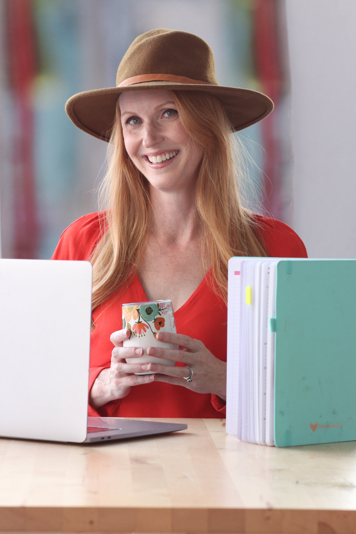 Woman with long red hair wearing a wide-brimmed hat and red top, smiling, holding a phone, sitting at a desk with a laptop and folders.