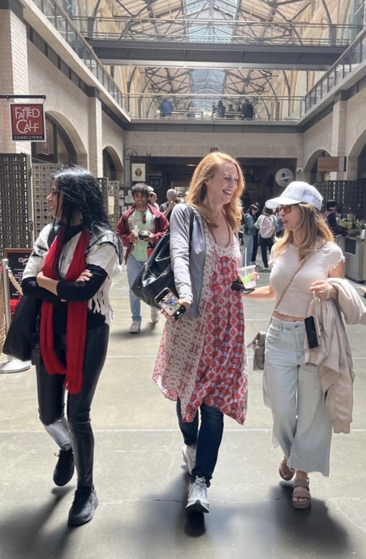 Three women walking in a shopping mall, with a glass ceiling and other shoppers in the background.