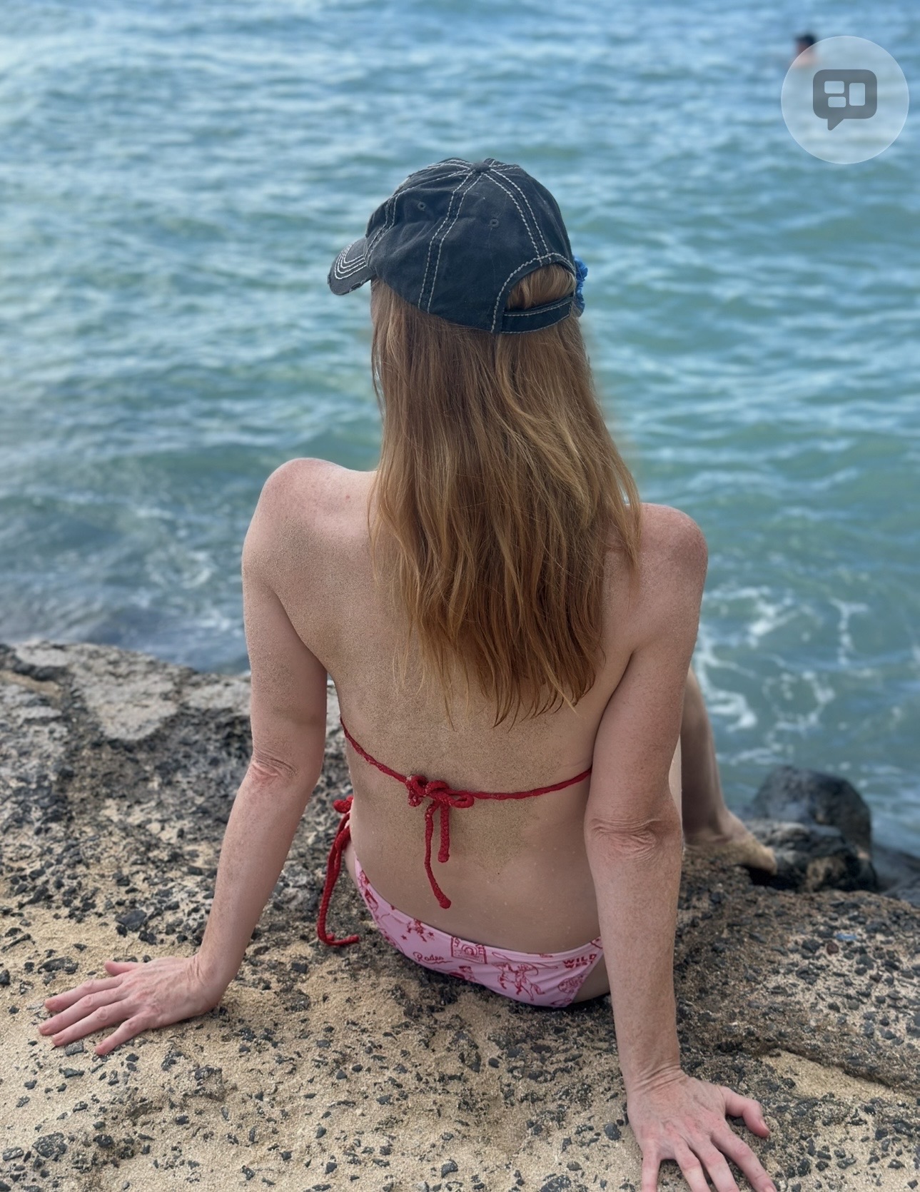 Person with long hair sitting on rocks by the water, wearing a cap and a bikini top, facing away from the camera.