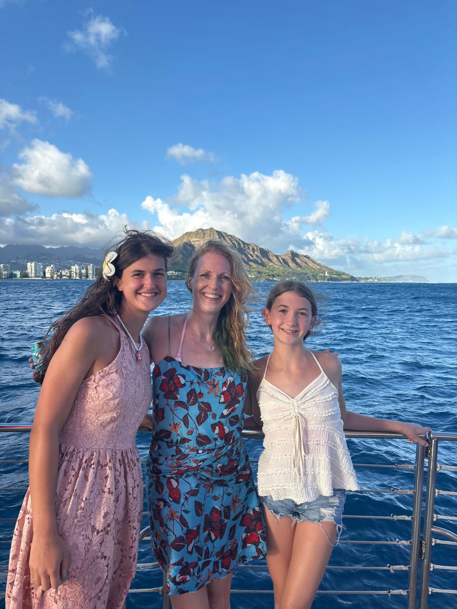 Three women stand on a boat railing by the water with a city skyline and mountains in the background.