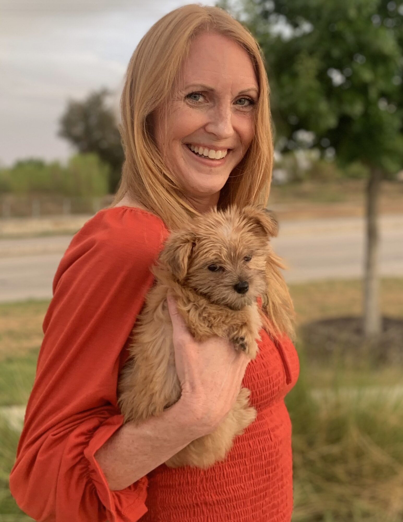 Woman with long red hair smiling and holding a small, fluffy puppy outdoors with trees and a fence in background.