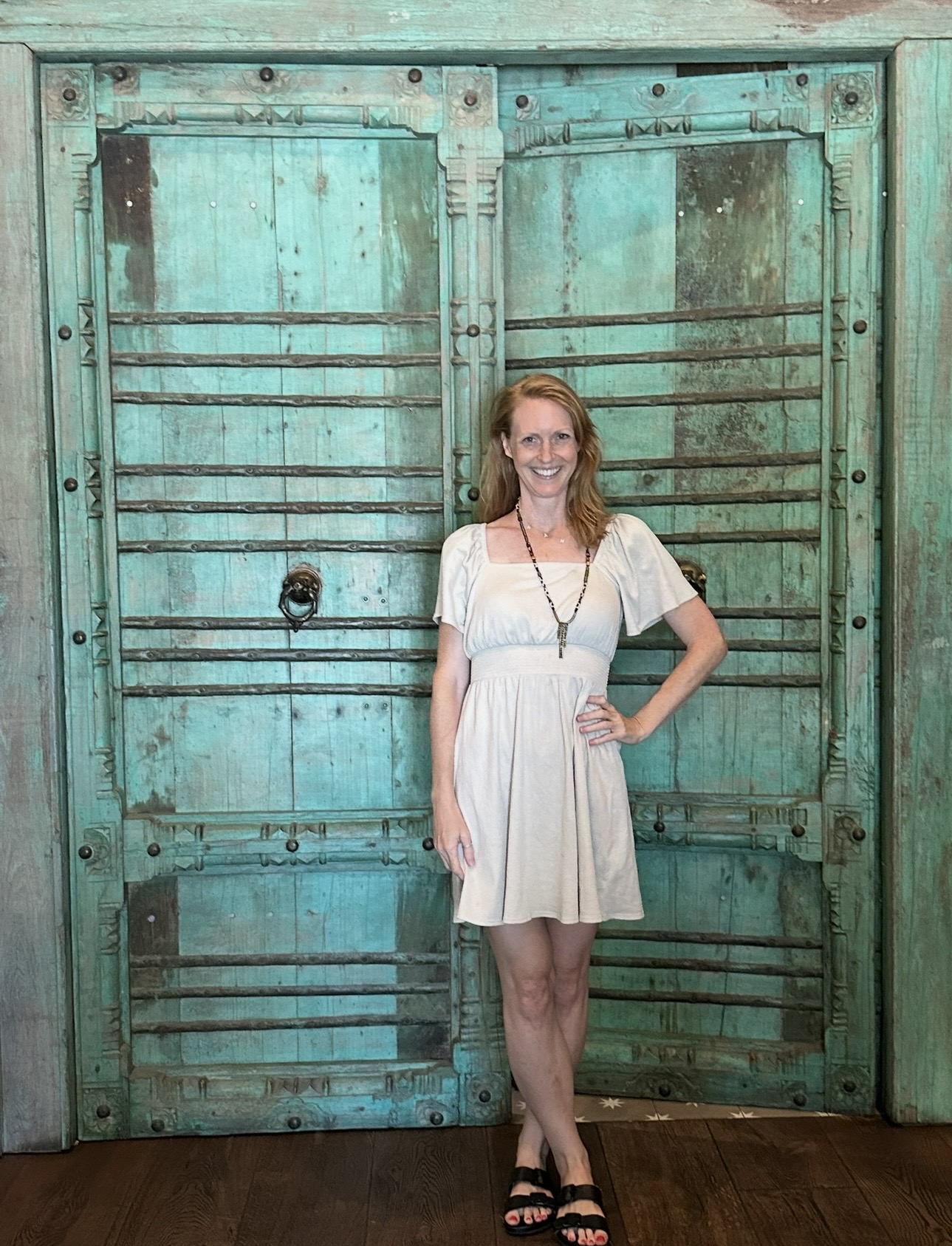 Woman in white dress standing in front of large turquoise wooden doors with metal bars, smiling, with one hand on her hip.