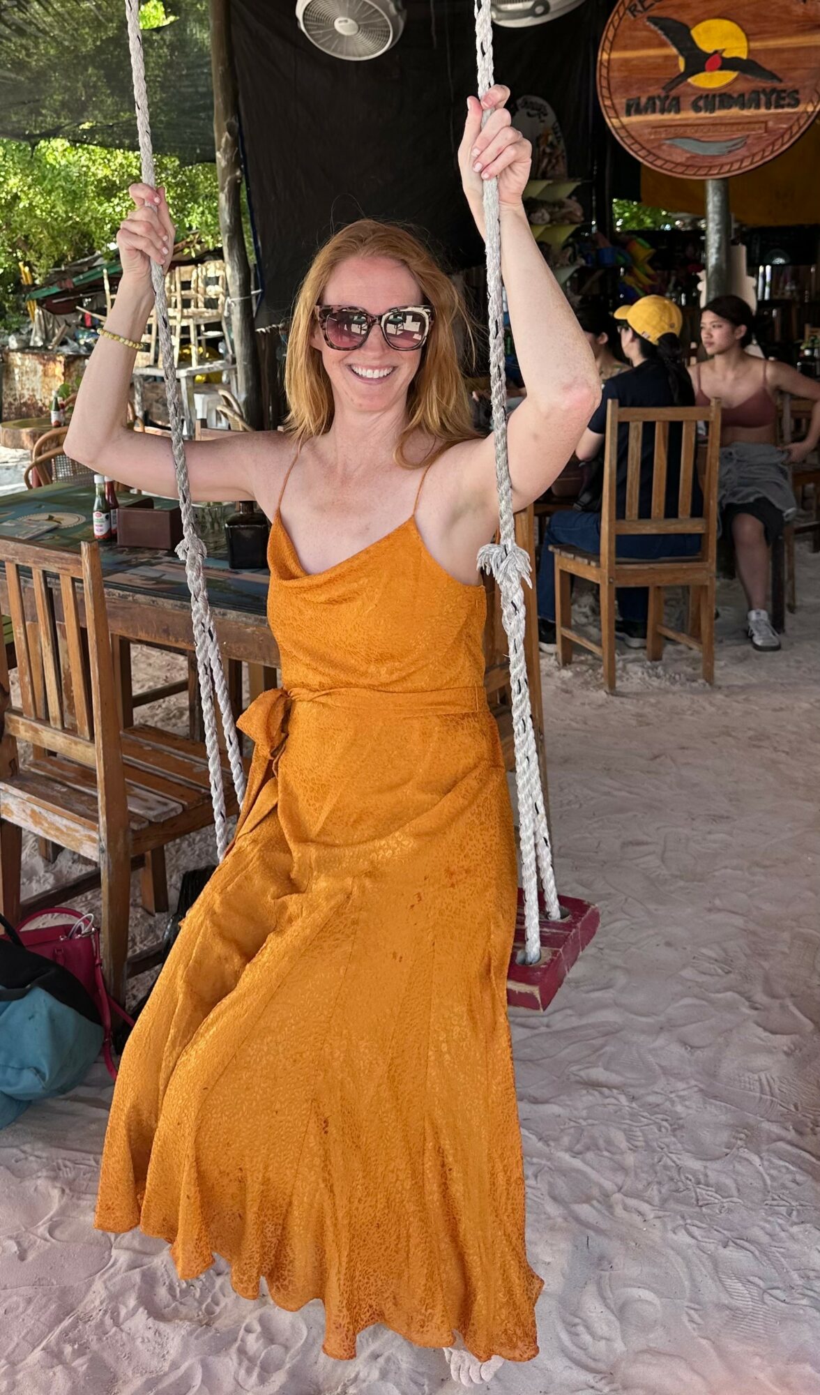 Woman in orange dress sitting on a swing at an outdoor venue, smiling, wearing sunglasses, with people and trees in background.