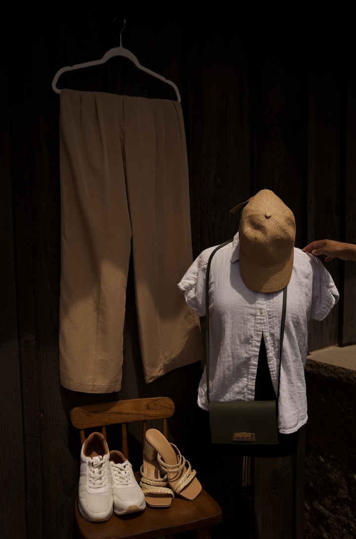 Child wearing a hat and white shirt, standing next to beige pants hanging on a hanger, with shoes on a small wooden stand.