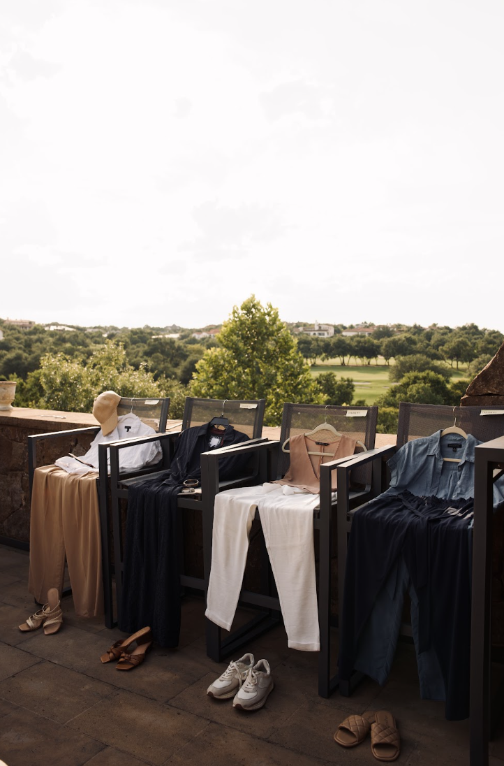 Clothes hanging on a railing outdoors with a landscape and trees in the background, shoes on the ground, and a clear sky.