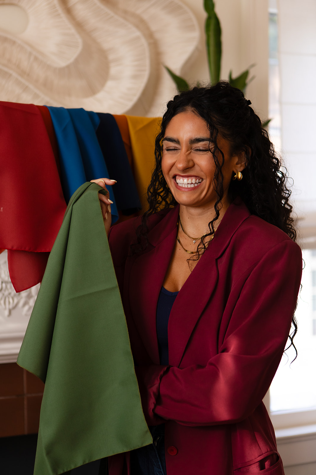 Woman with curly hair smiling, holding green fabric, colorful fabrics hanging behind her, indoors with plants and natural light.