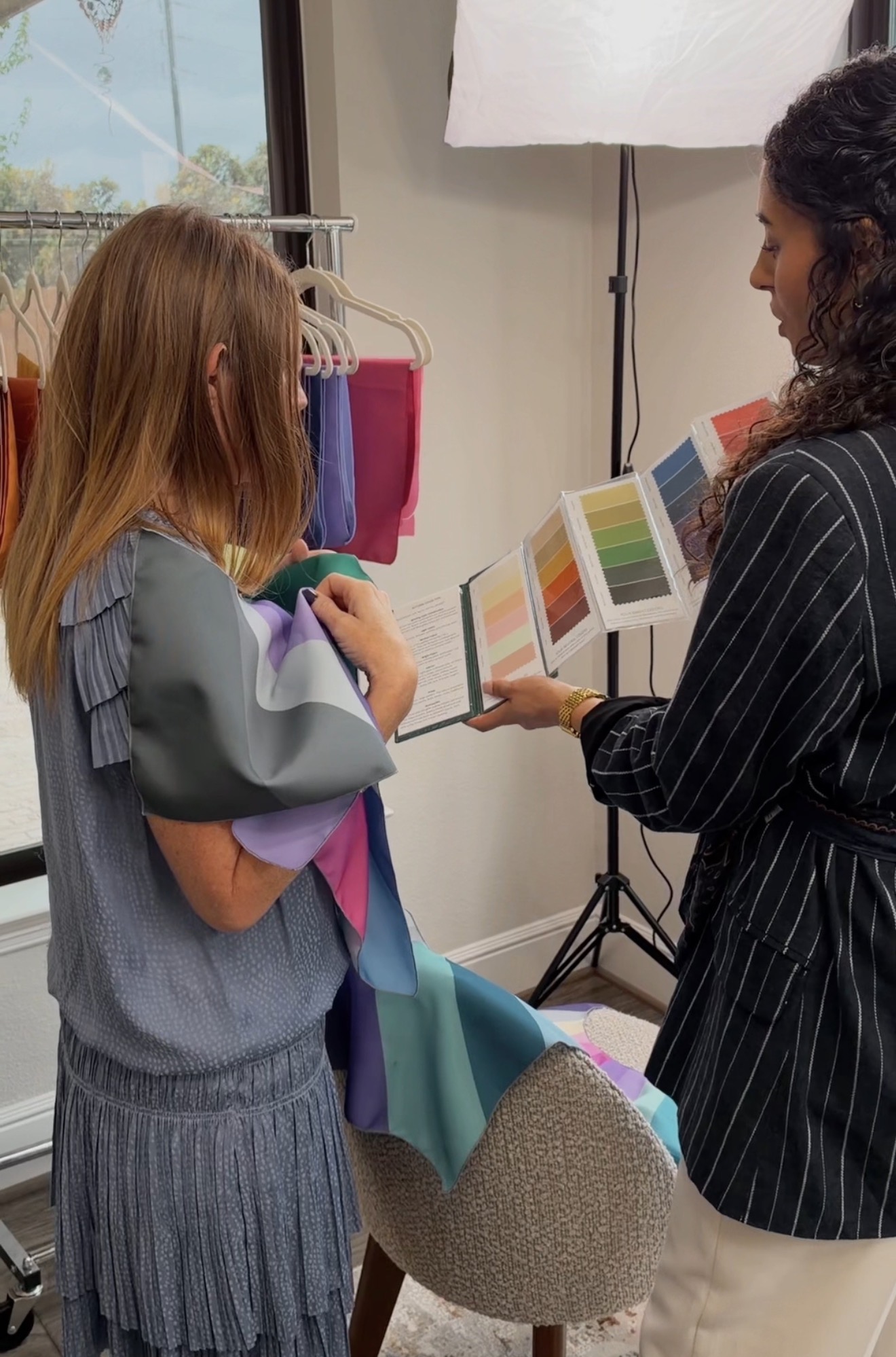 Two women look at fabric samples in a room with a window and clothing rack, discussing colors and textures.