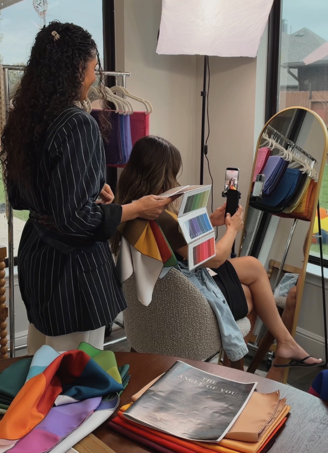 Two women in a room, one sitting and taking a selfie, the other standing nearby, with clothing and a mirror visible.