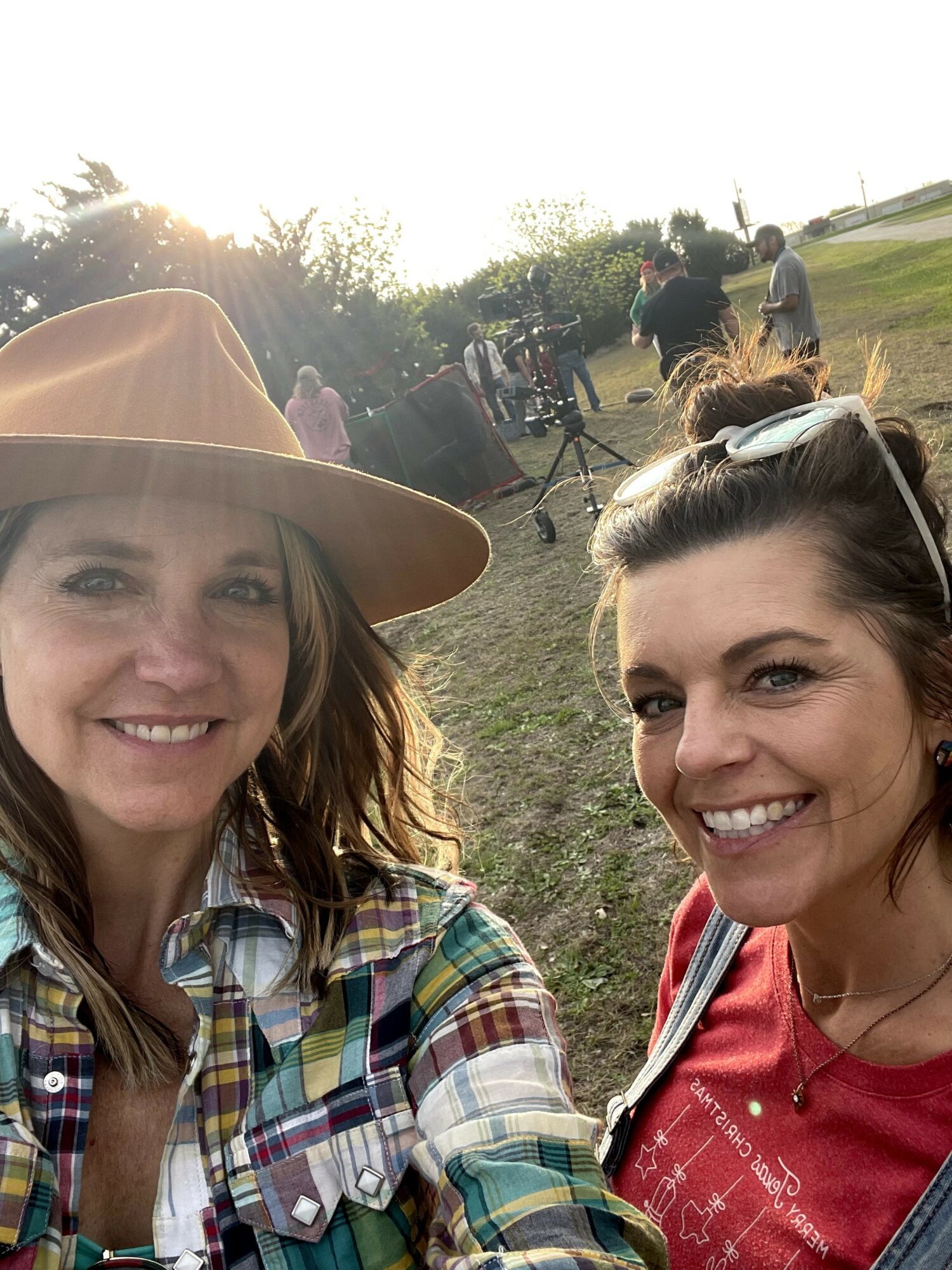 Two women smiling outdoors with people and trees in the background, sunlight shining behind them.