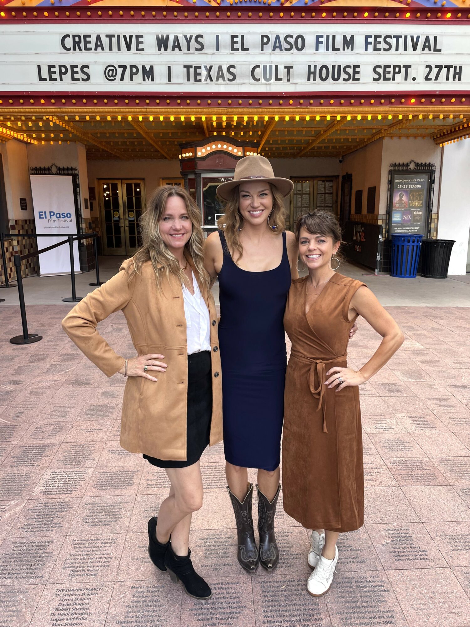 Three women standing together in front of a theater entrance, smiling, with a marquee above them displaying event details.