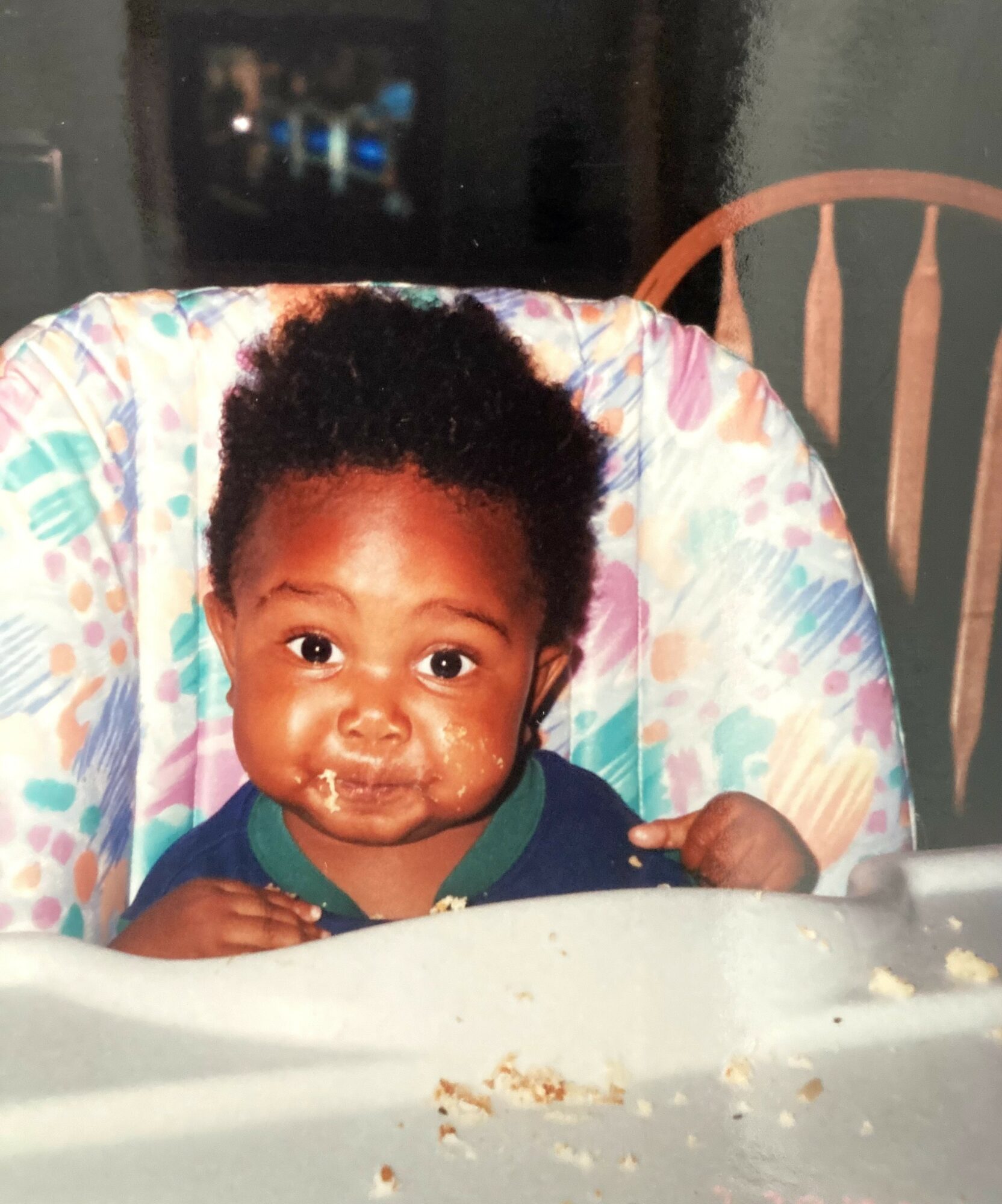 Baby with curly hair sitting in a high chair, looking at the camera, with food on face and hands, in a dining area.