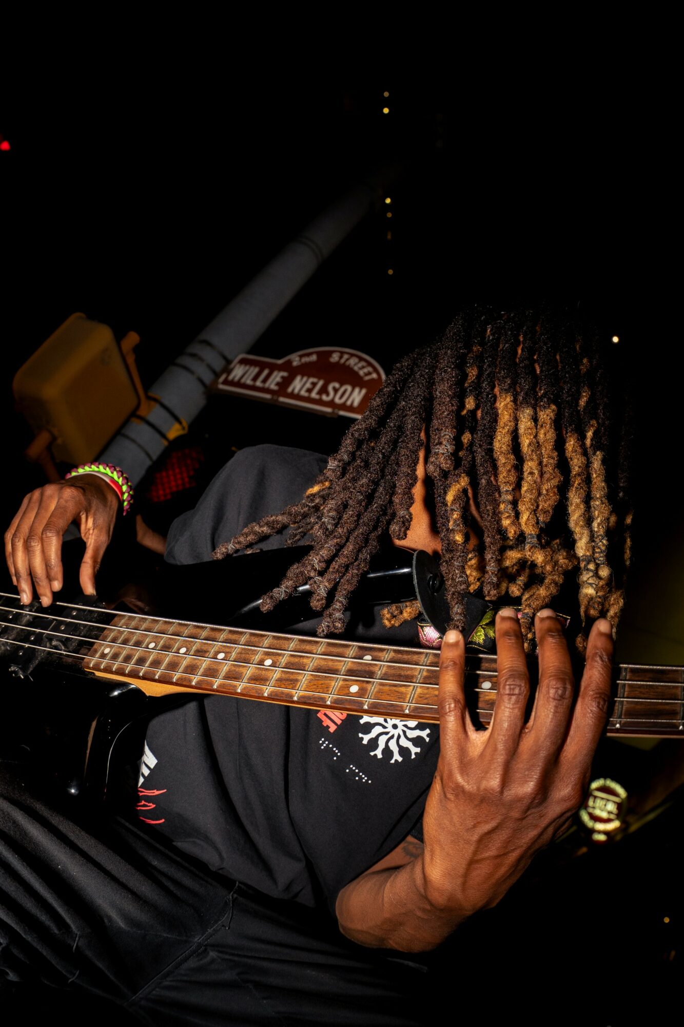 Person with dreadlocks playing guitar, dark background, visible hand and part of guitar neck, wearing rings and bracelets.