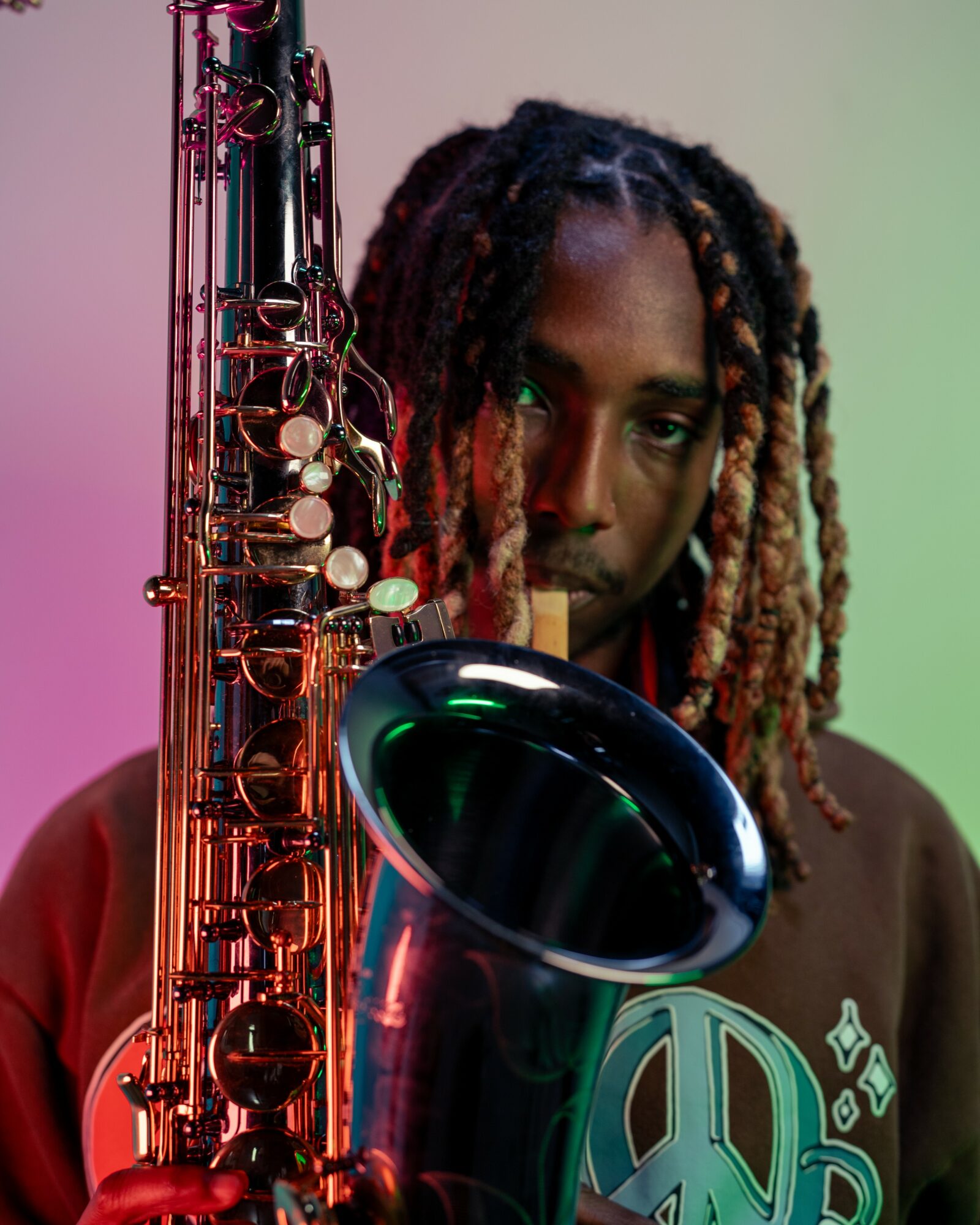 Person with dreadlocks holding a saxophone, looking at the camera, with colorful lighting background.