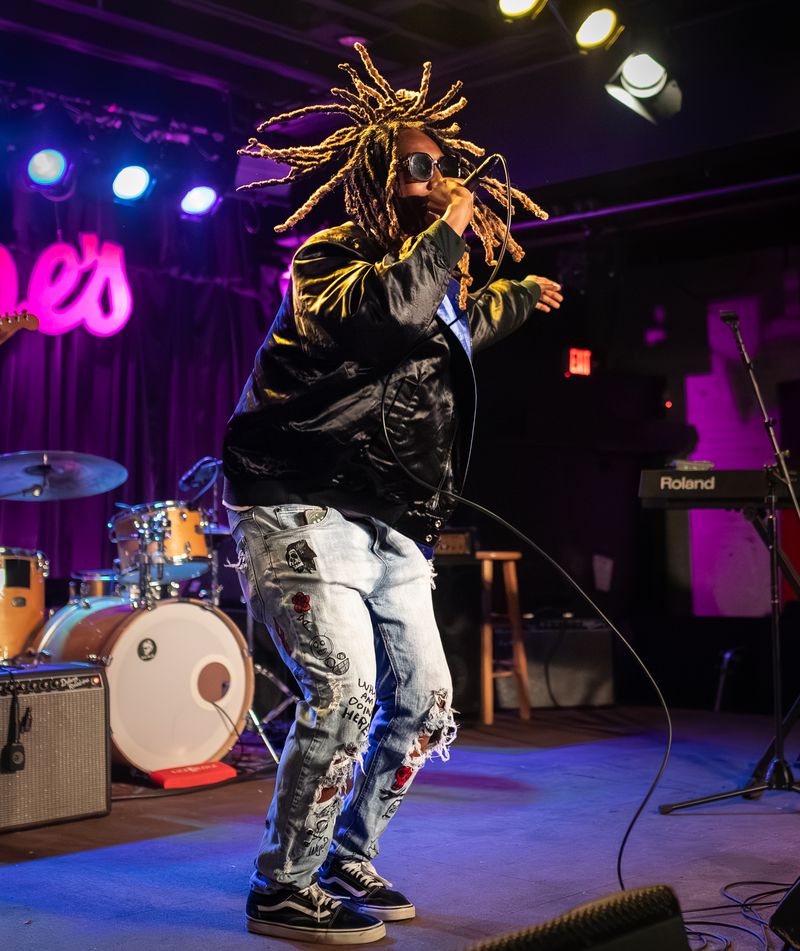 Performer with dreadlocks, sunglasses, black jacket, and ripped jeans singing on stage with musical instruments behind.