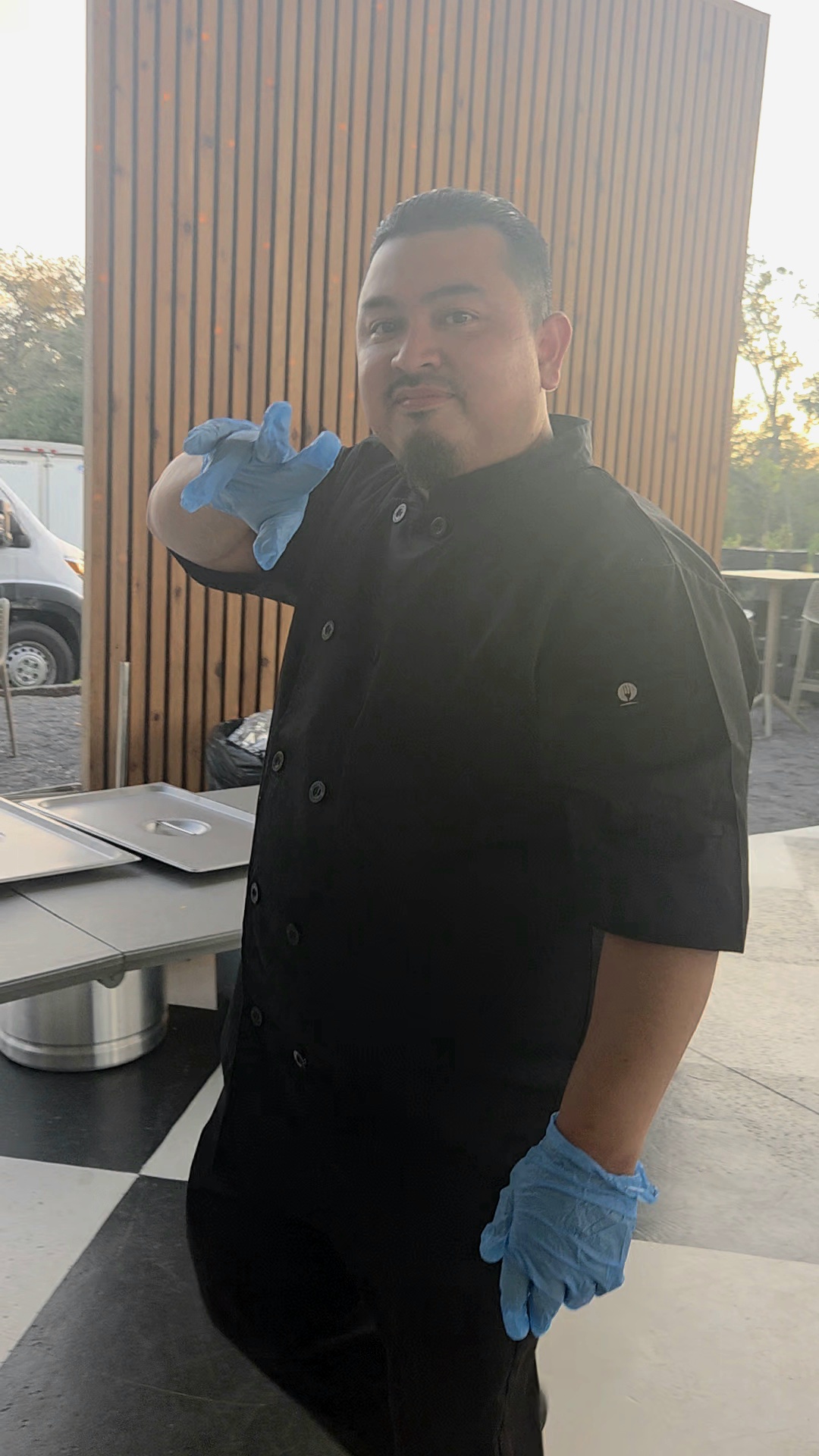 Person wearing black uniform and blue gloves, standing indoors near wooden wall, making a gesture with hand.