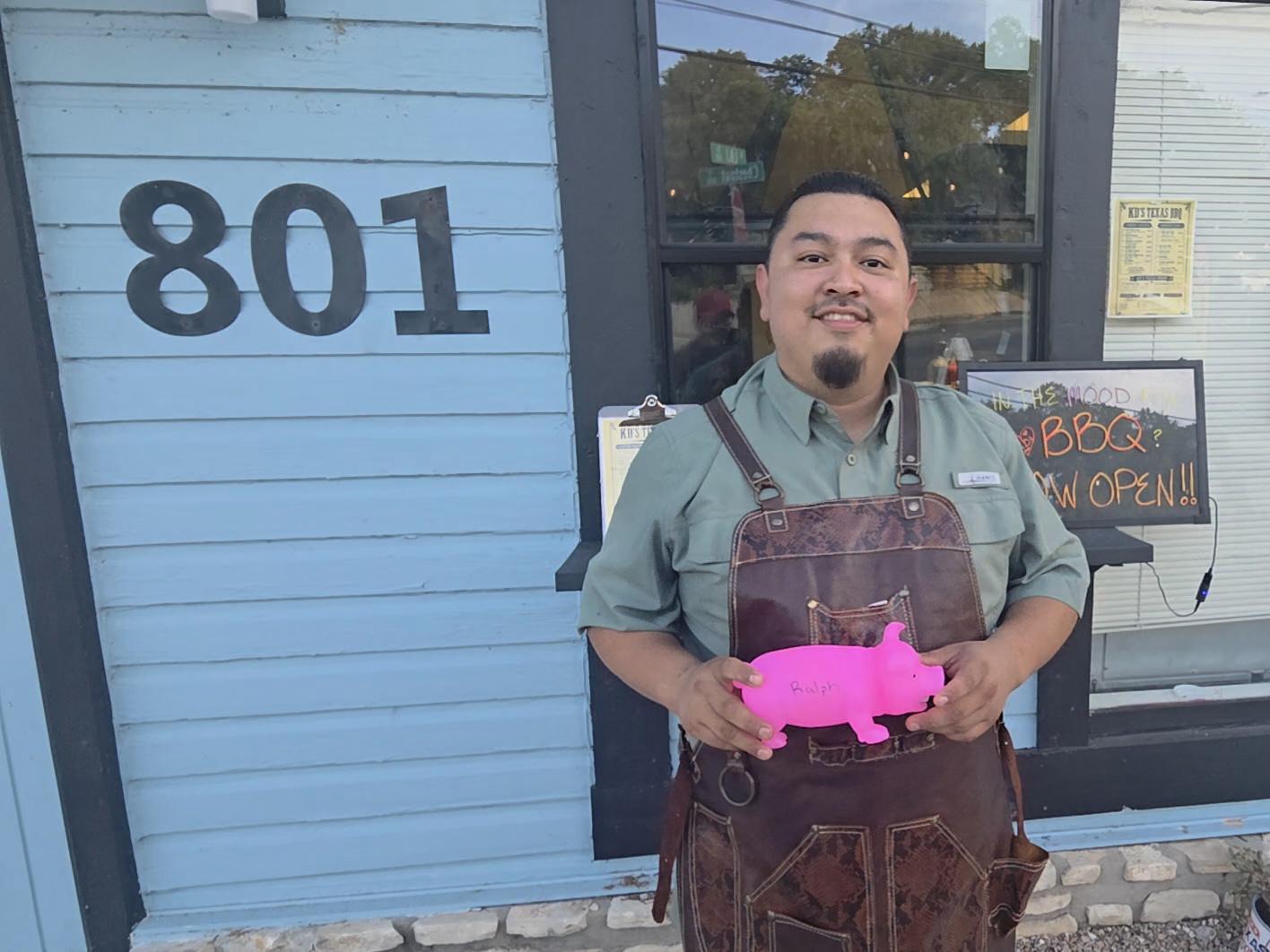 Man standing outside a building with the number 801, holding a pink pig-shaped object, smiling.