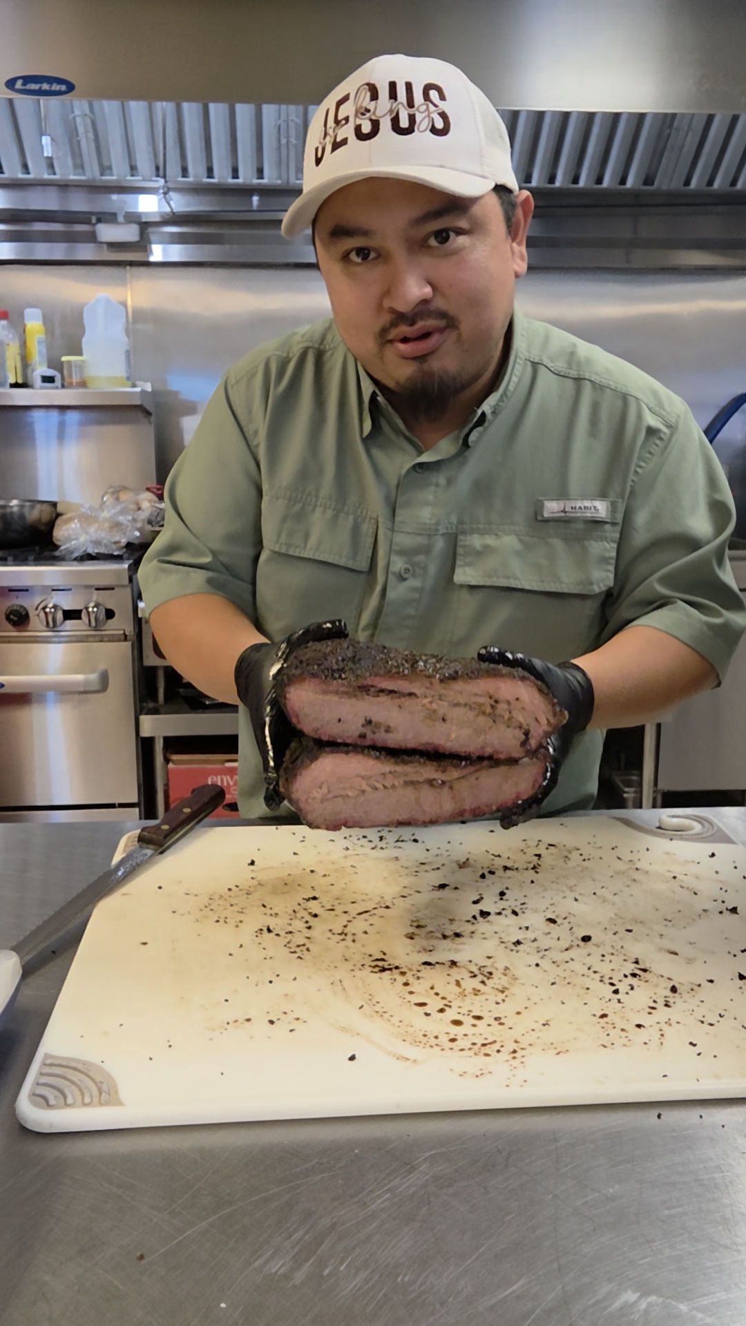 Man wearing a white cap and green shirt holding a large cooked meat cut in a kitchen.