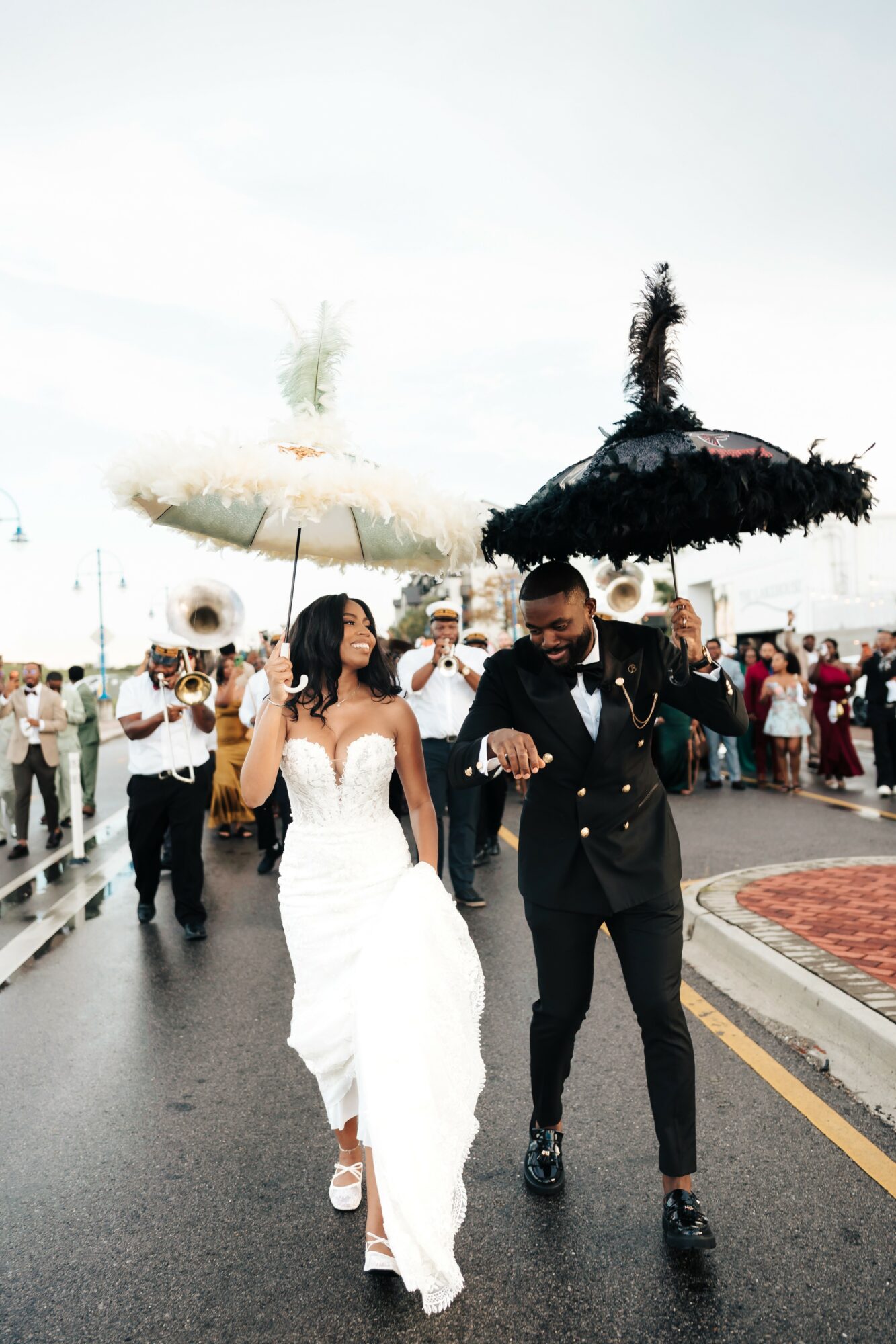 Couple in wedding attire walking on a street during a celebration, holding decorated umbrellas, with a crowd in the background.