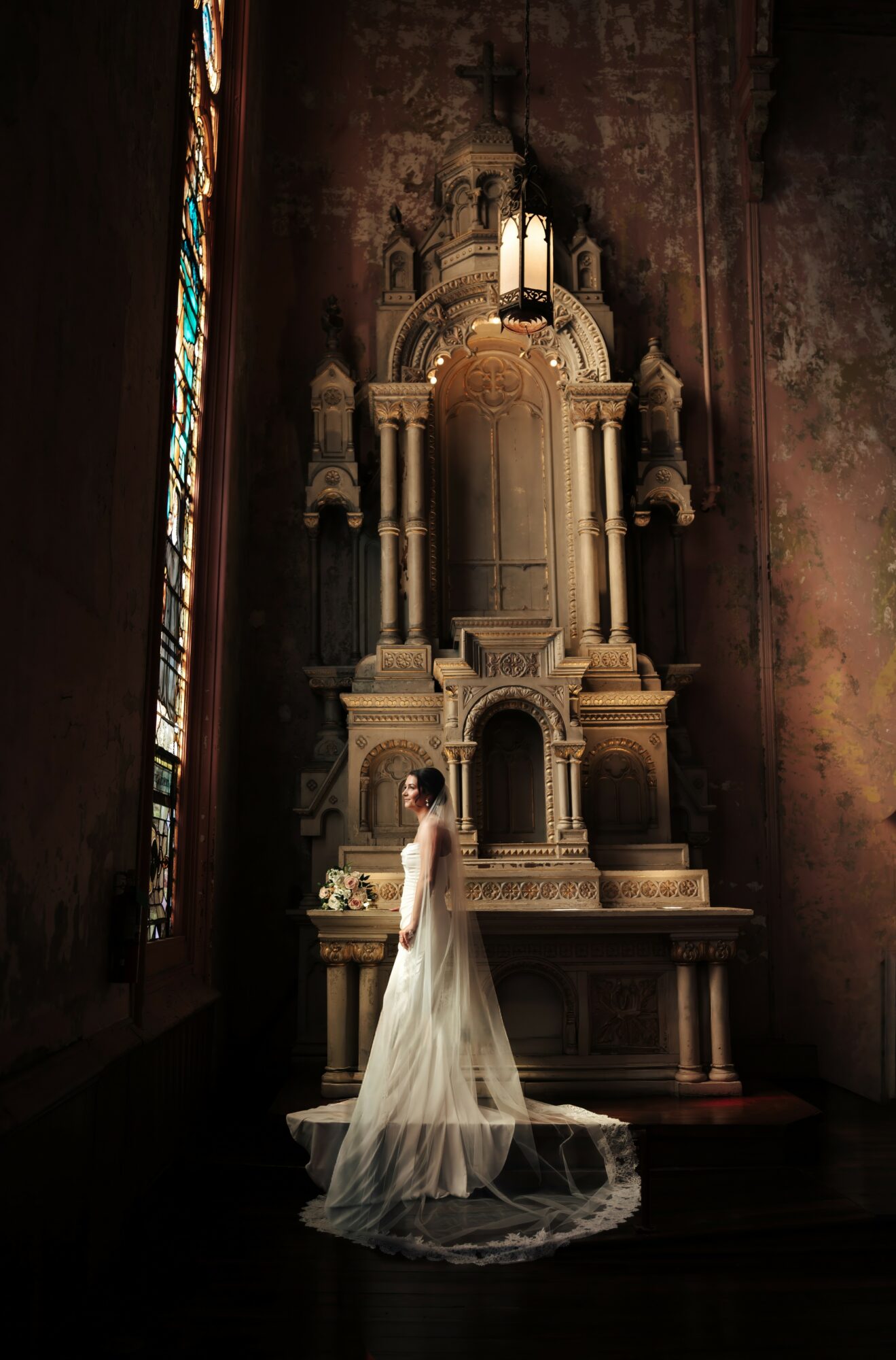 A woman in a white dress stands in front of an ornate altar inside a dimly lit church with stained glass window.