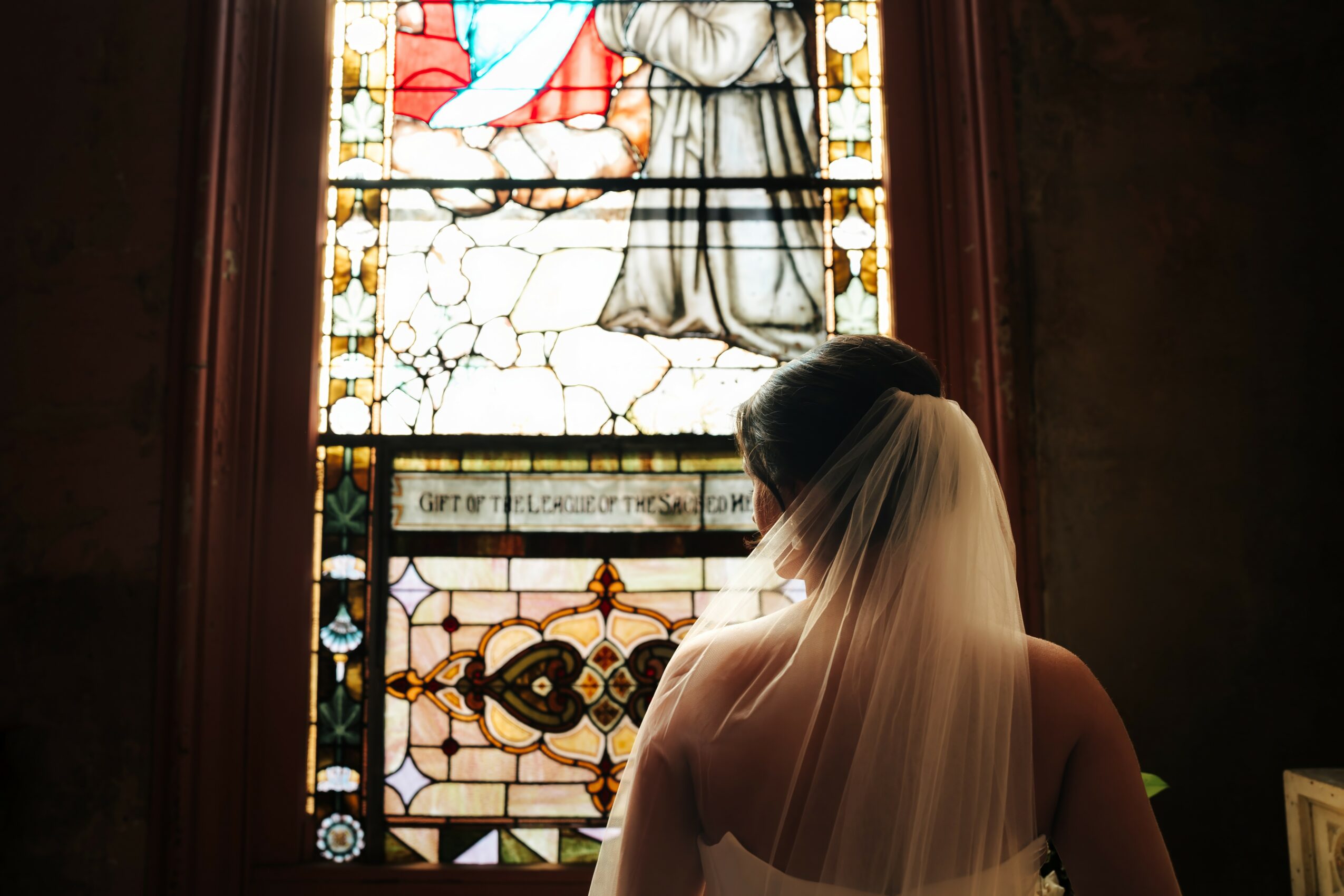 Bride with veil facing stained glass window inside a church, with dark interior surroundings.