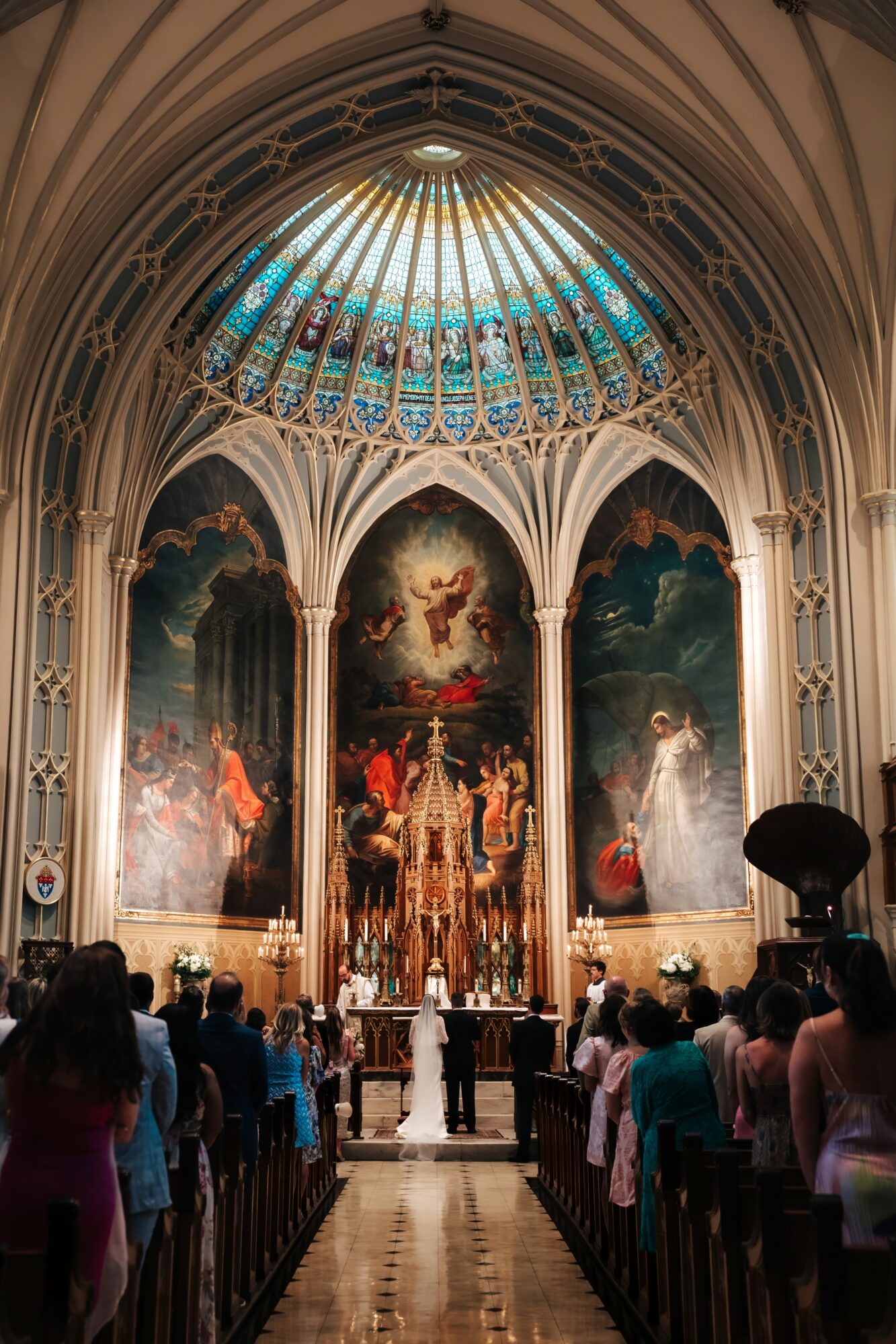 Interior of a church with a stained glass dome, large religious paintings, and people attending a service.