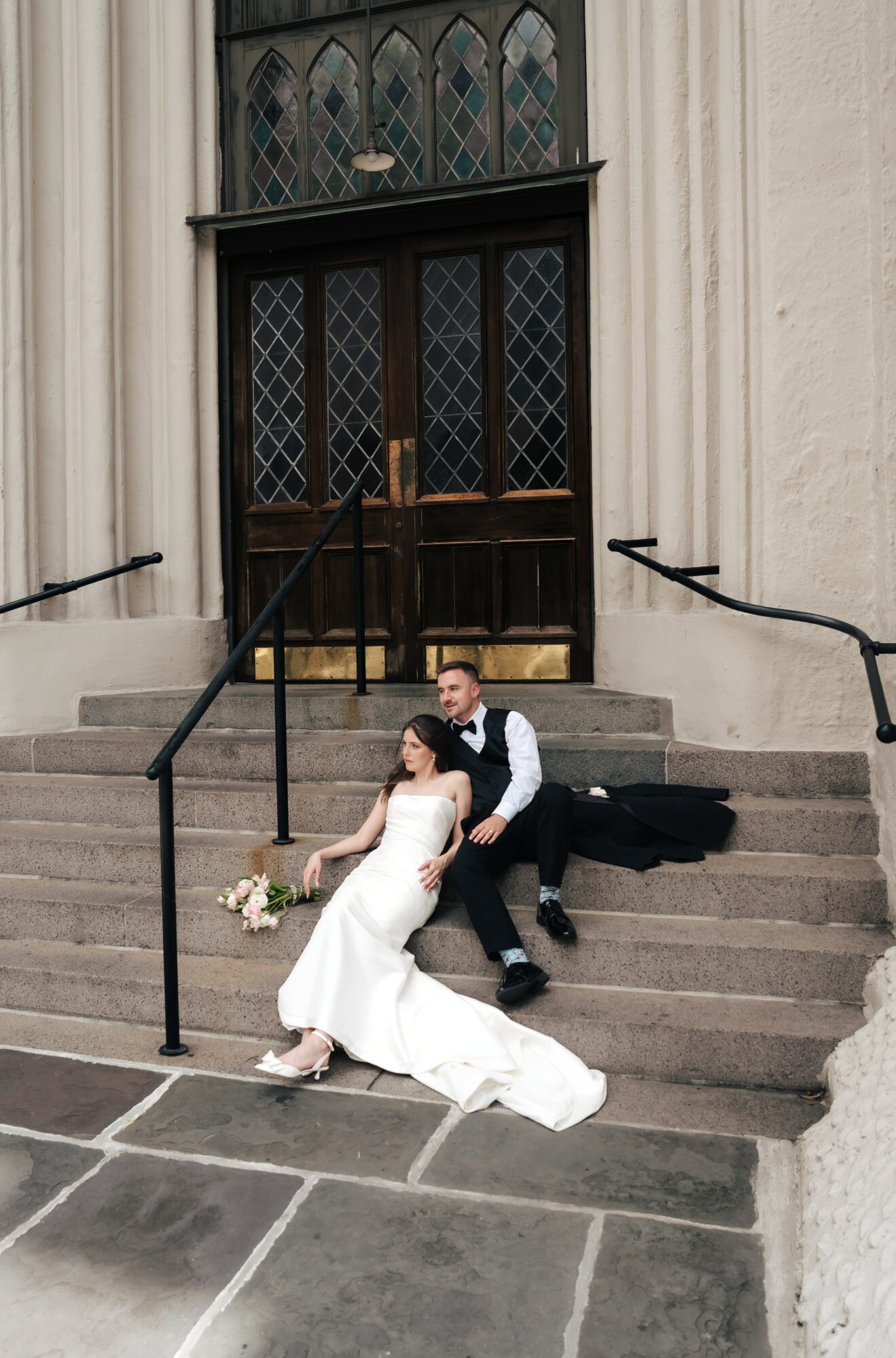 A bride and groom sit on church steps, the bride holding a bouquet, in front of a large wooden door with stained glass.