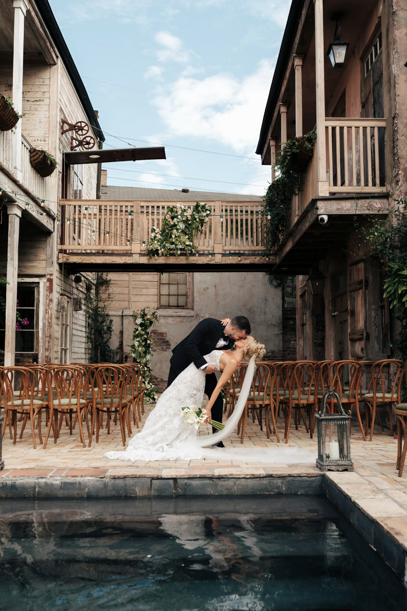 Couple kissing by a pool in an outdoor courtyard with wooden buildings and chairs, sky visible above.