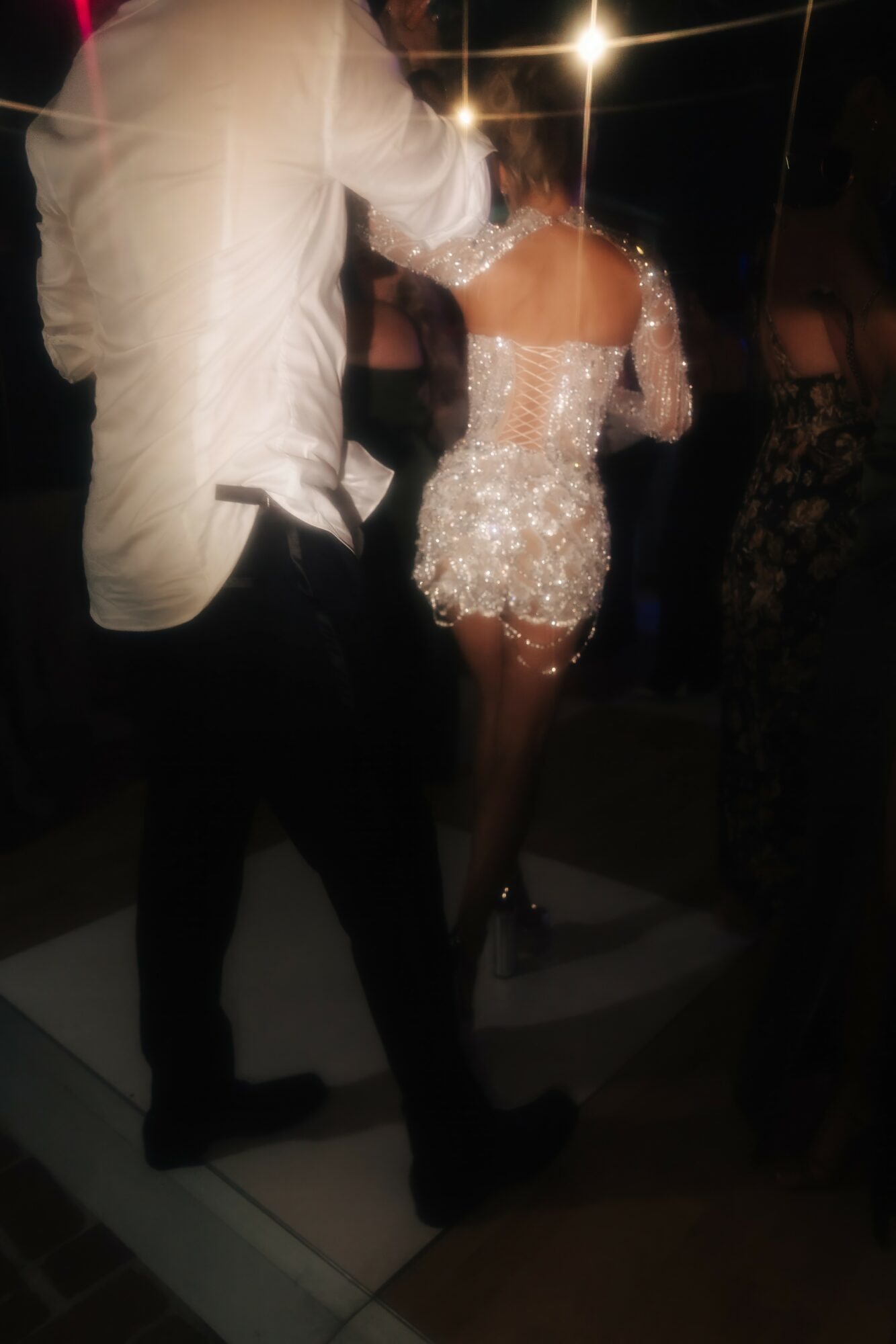Person in a white shirt and black pants dancing with a woman in a sparkly dress, in a dimly lit setting.