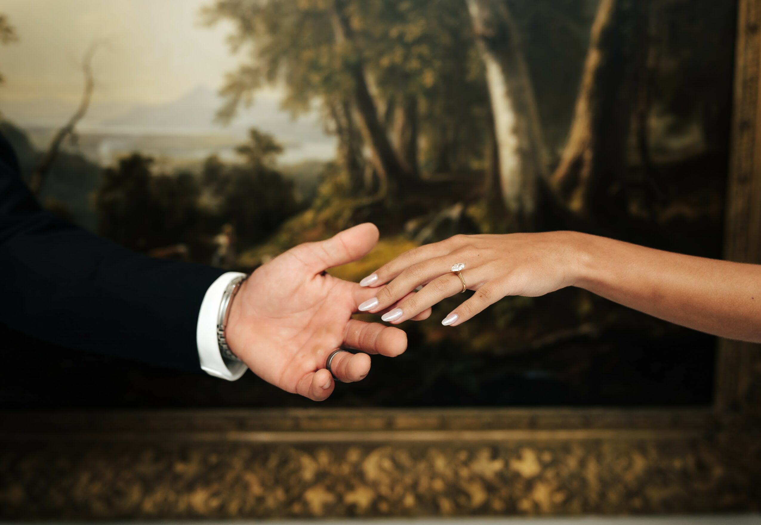 Two hands reaching towards each other, one with a wedding ring, in front of a landscape painting.