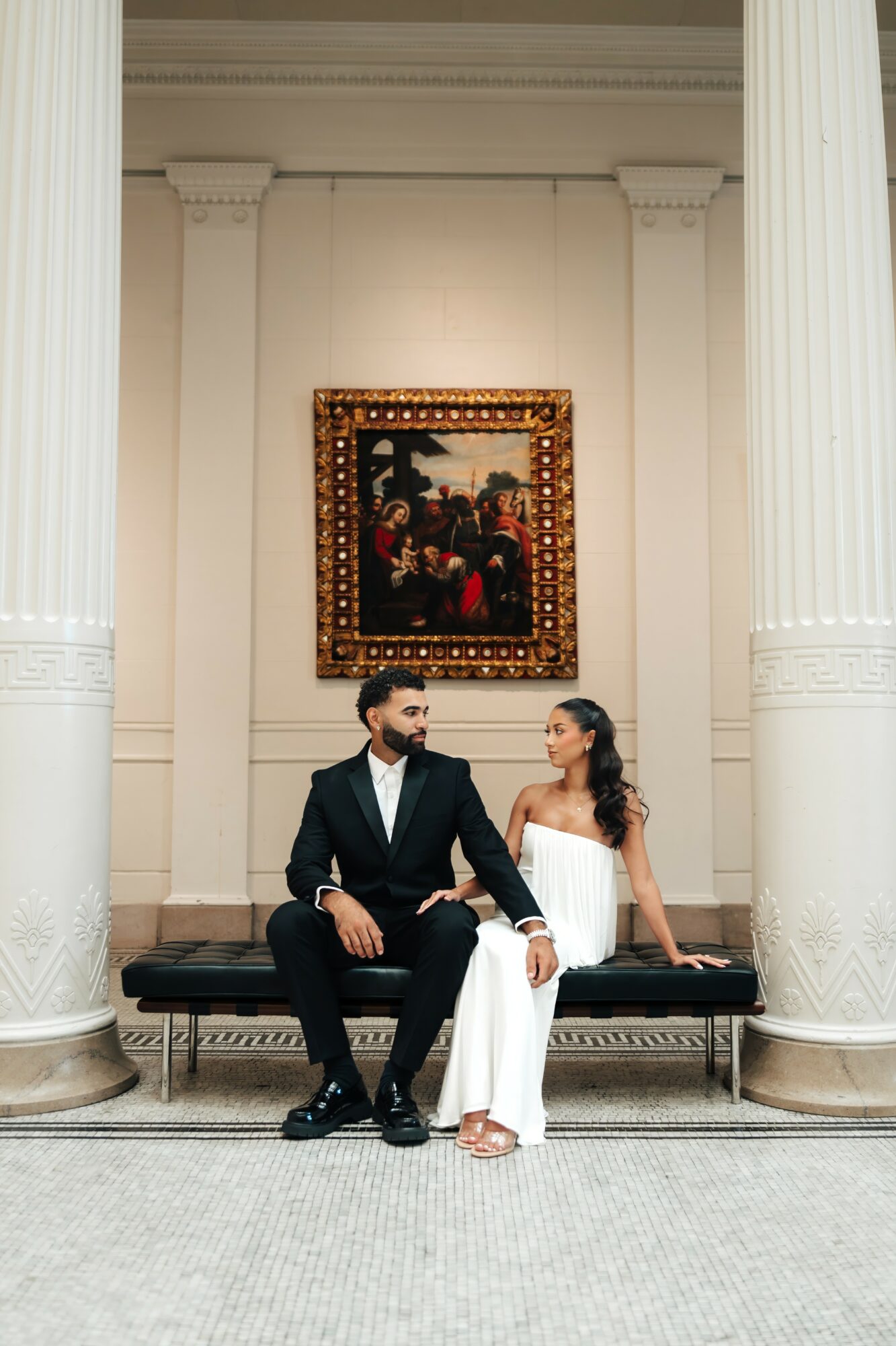 A man and woman sit on a bench in a grand hall with tall columns and a framed painting behind them.