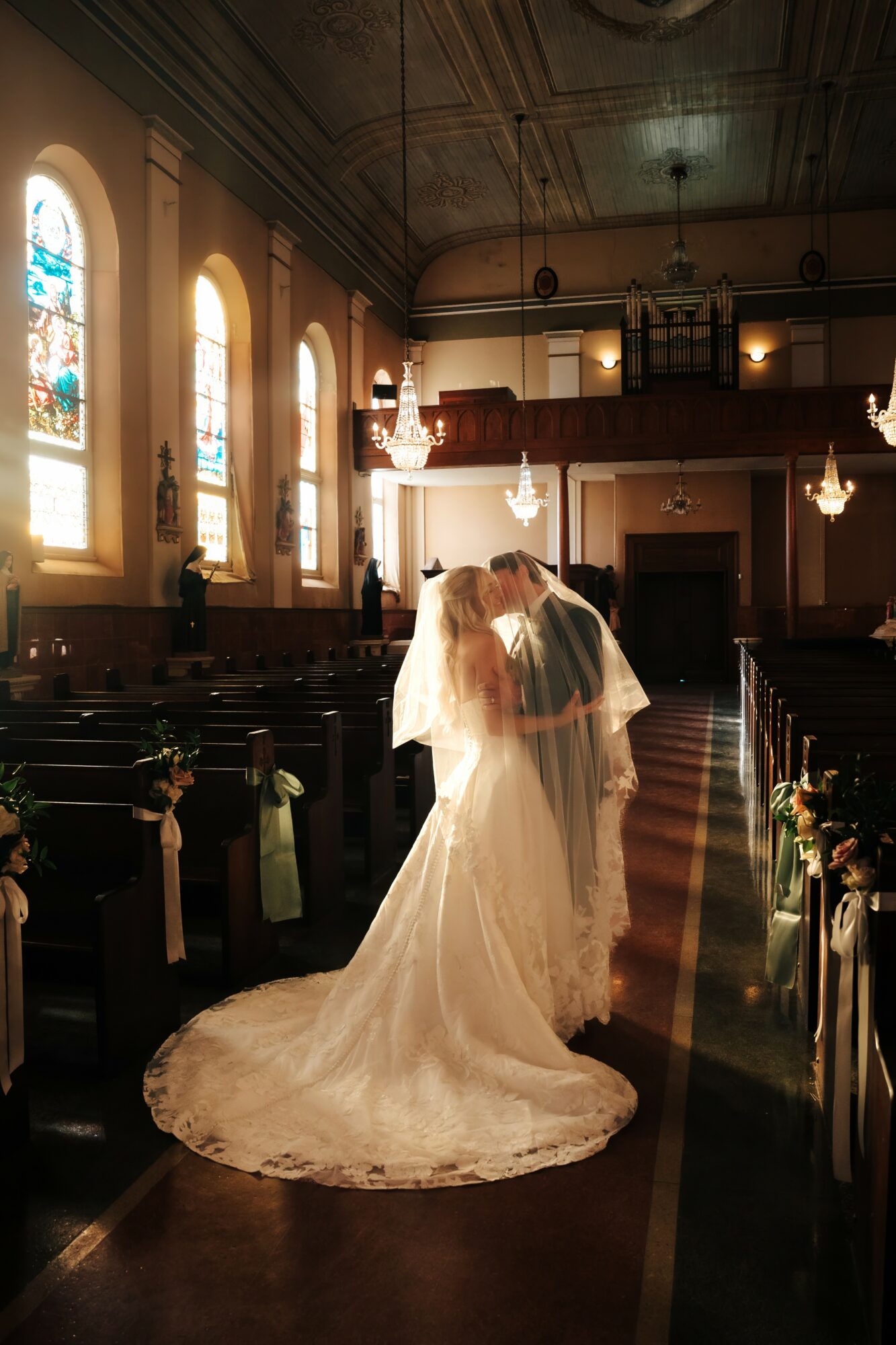 Bride and groom kissing inside a church with stained glass windows, pews, and chandeliers, bride in a long white gown with train.