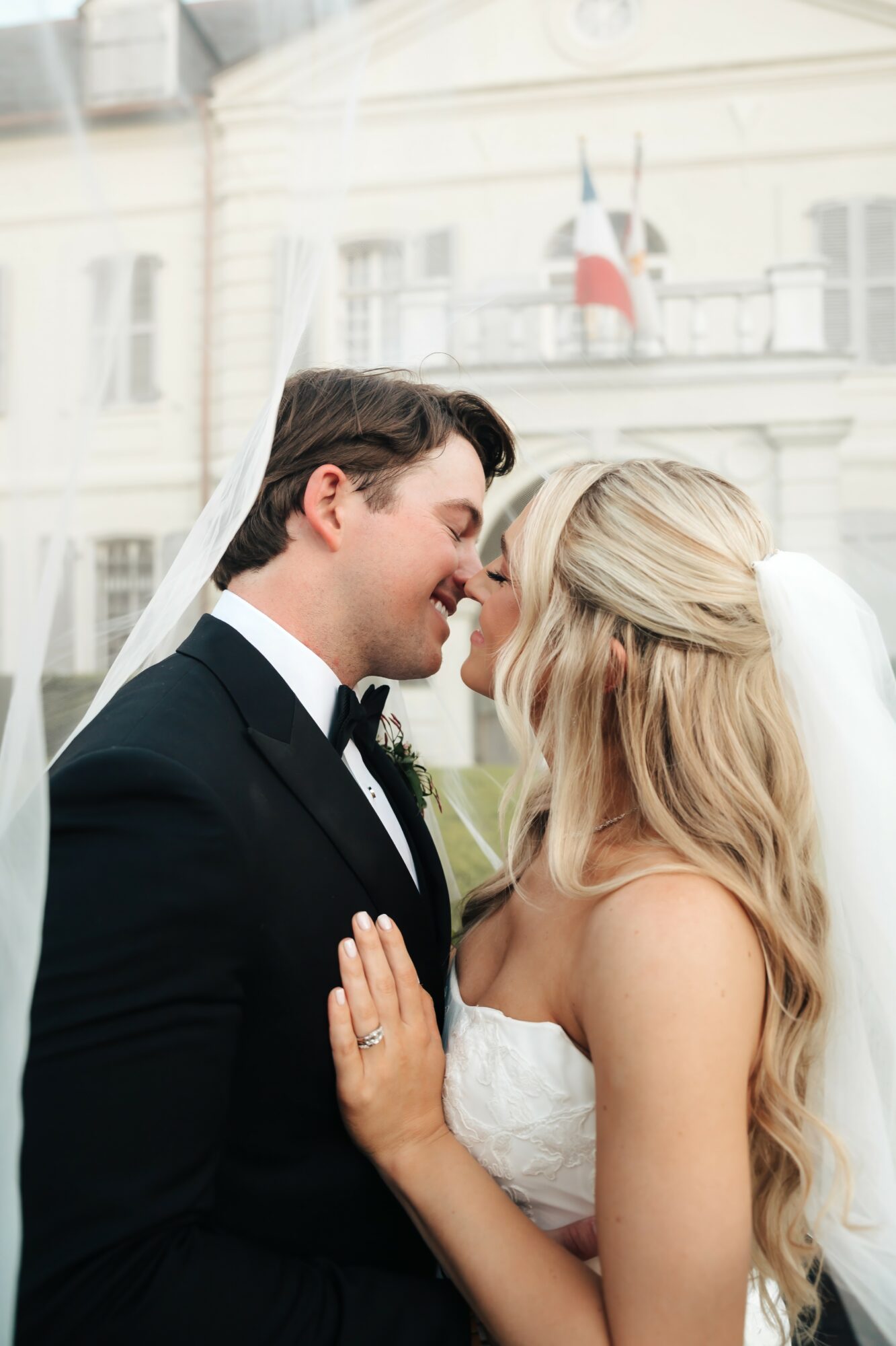 A bride and groom face each other closely, smiling, with a building and French flag in the background.