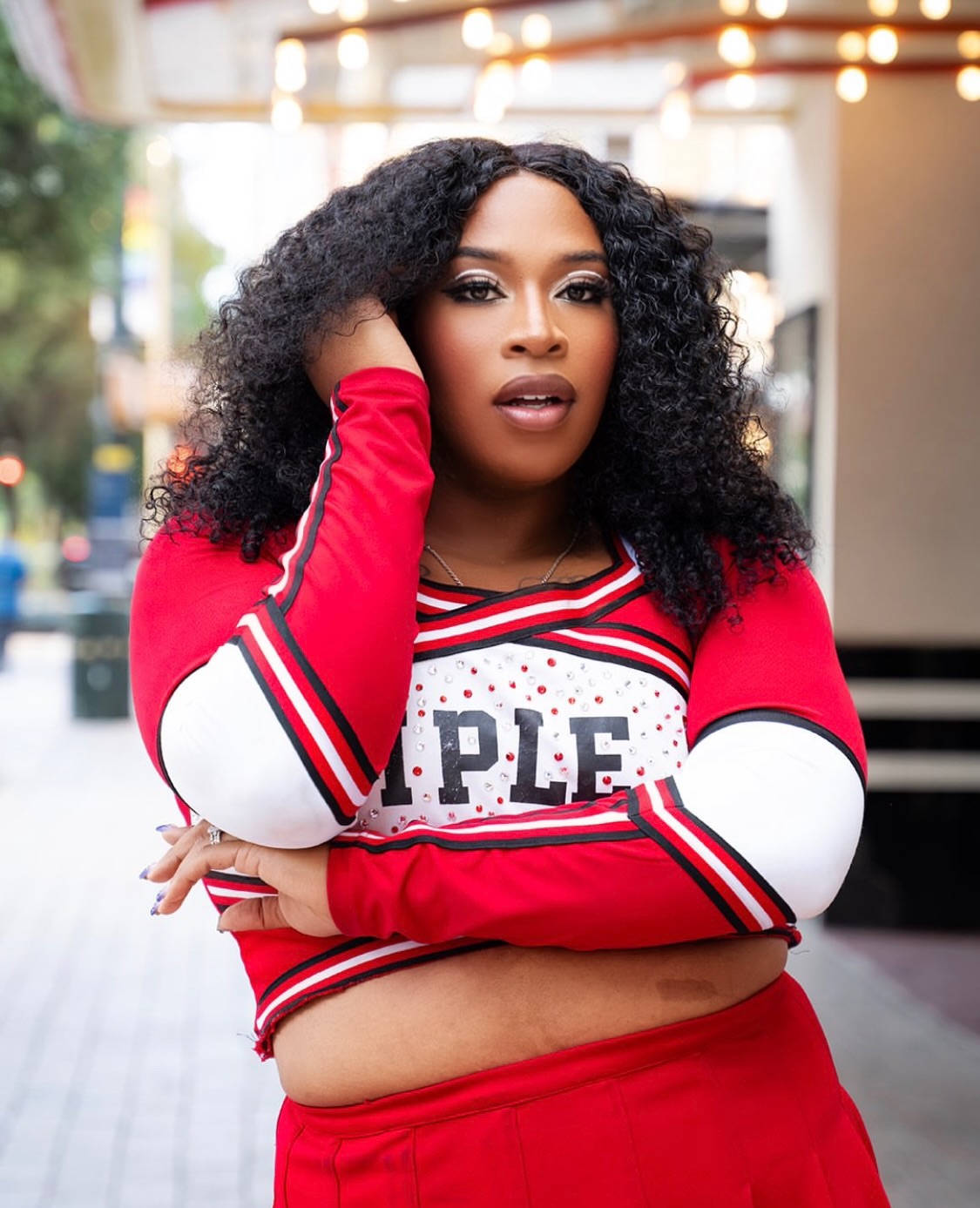 Woman with curly hair wearing a red and white cheerleader-style top, standing outdoors with blurred background.