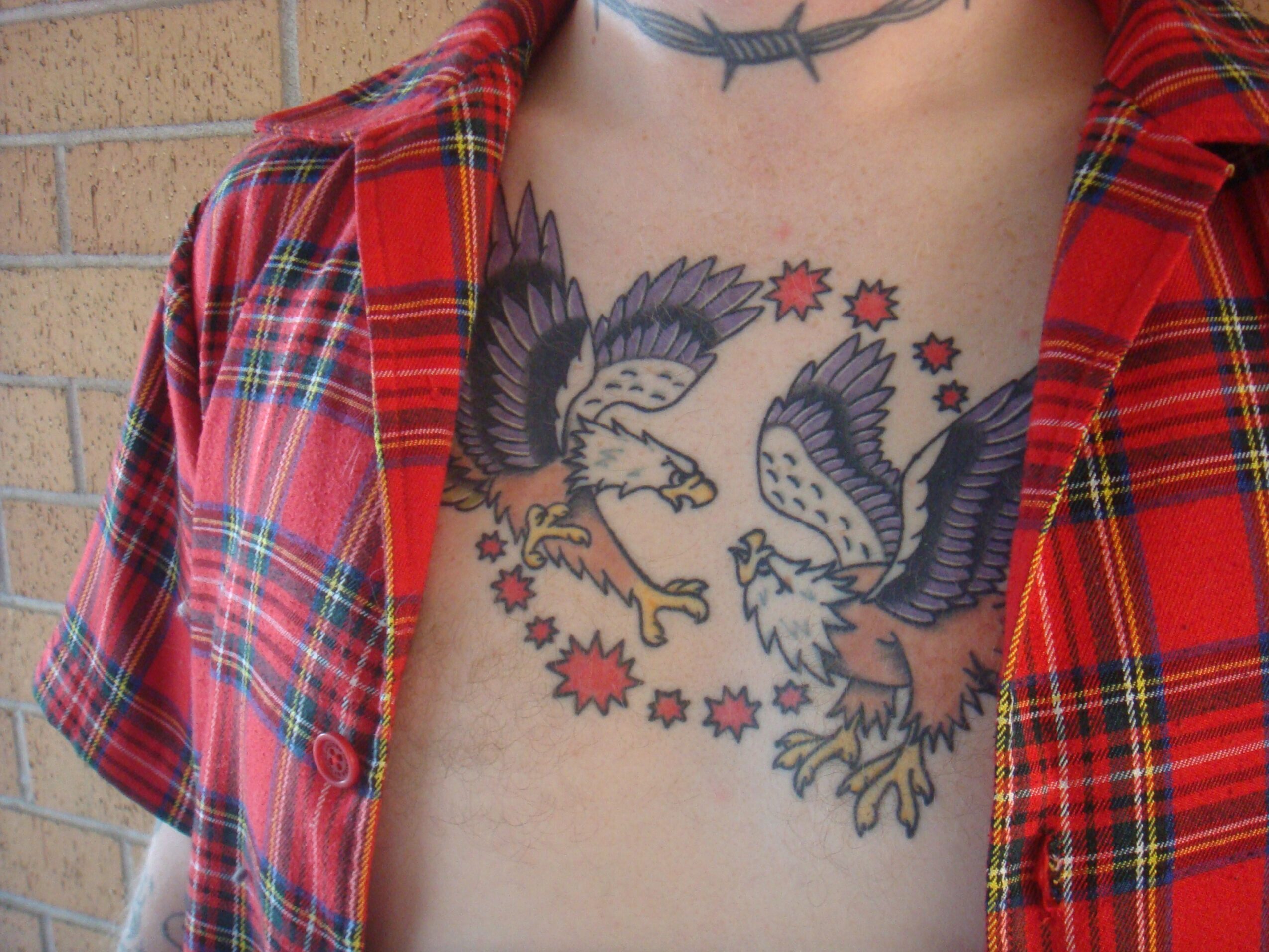 Tattoo of two eagles facing each other with stars around them on person's chest, partially covered by red plaid shirt.