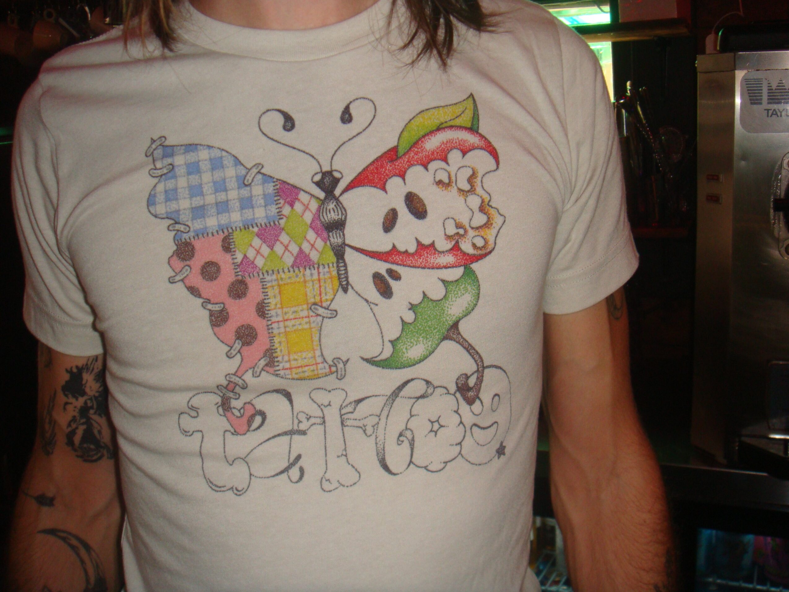 Person wearing a white T-shirt with a colorful butterfly and cupcake design, tattoos visible on arms.