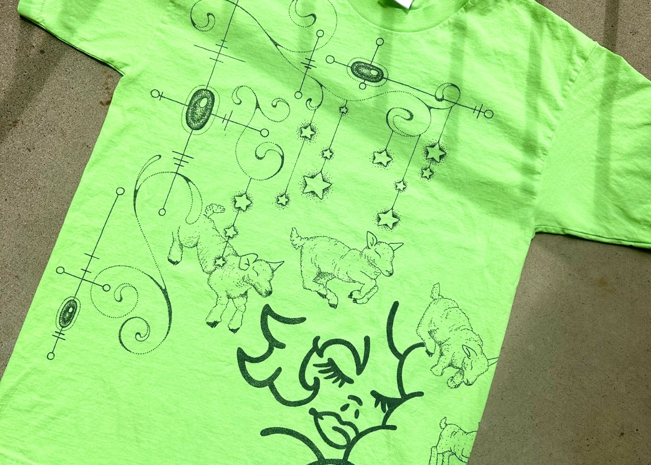 Green T-shirt with black line drawings of animals, stars, and abstract designs, laid on a gray surface.