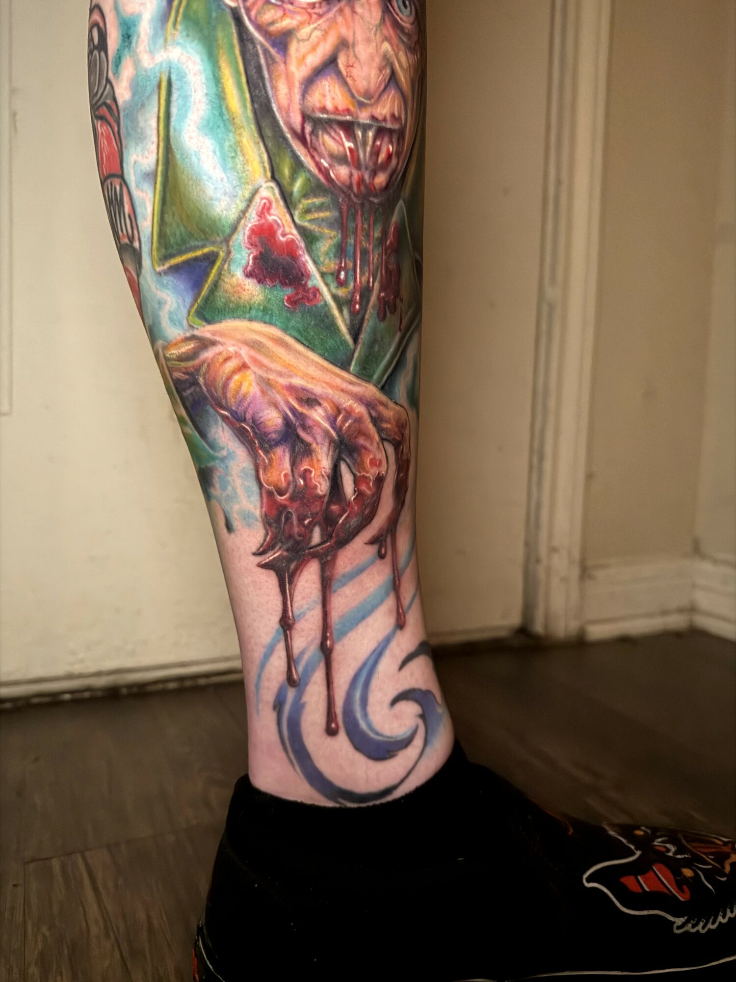 Tattooed leg featuring a colorful, detailed design with a face, hand, and abstract elements, against a beige background.